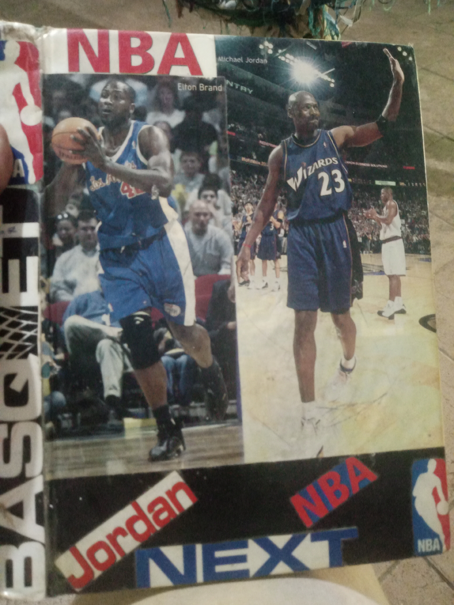 The image is a close-up, angled shot of the cover of what appears to be a sports magazine or book featuring NBA basketball players. The cover is divided into two main panels. The left panel shows a player identified as Elton Brand in a blue and white uniform, holding a basketball. The right panel features Michael Jordan in a blue Wizards uniform with the number 23, his arm raised. The background of both panels shows basketball courts, crowds, and other players.
At the bottom of the cover, text overlays the images. "Jordan" is written diagonally in red letters on a white background. Below that, "NEXT" is written in large blue letters with a white outline. The NBA logo, a silhouette of a player dribbling a ball, is visible on the far right, next to the word "NBA" in red letters on a blue background. Vertical text reading "BAS" is visible on the far left edge. The cover exhibits signs of wear and tear, including creases and scratches.
This image does not contain any information that allows for the determination of its location in Santa Ana, Venezuela, or any other city. The scene depicted is a professional sports event, not a specific urban location.