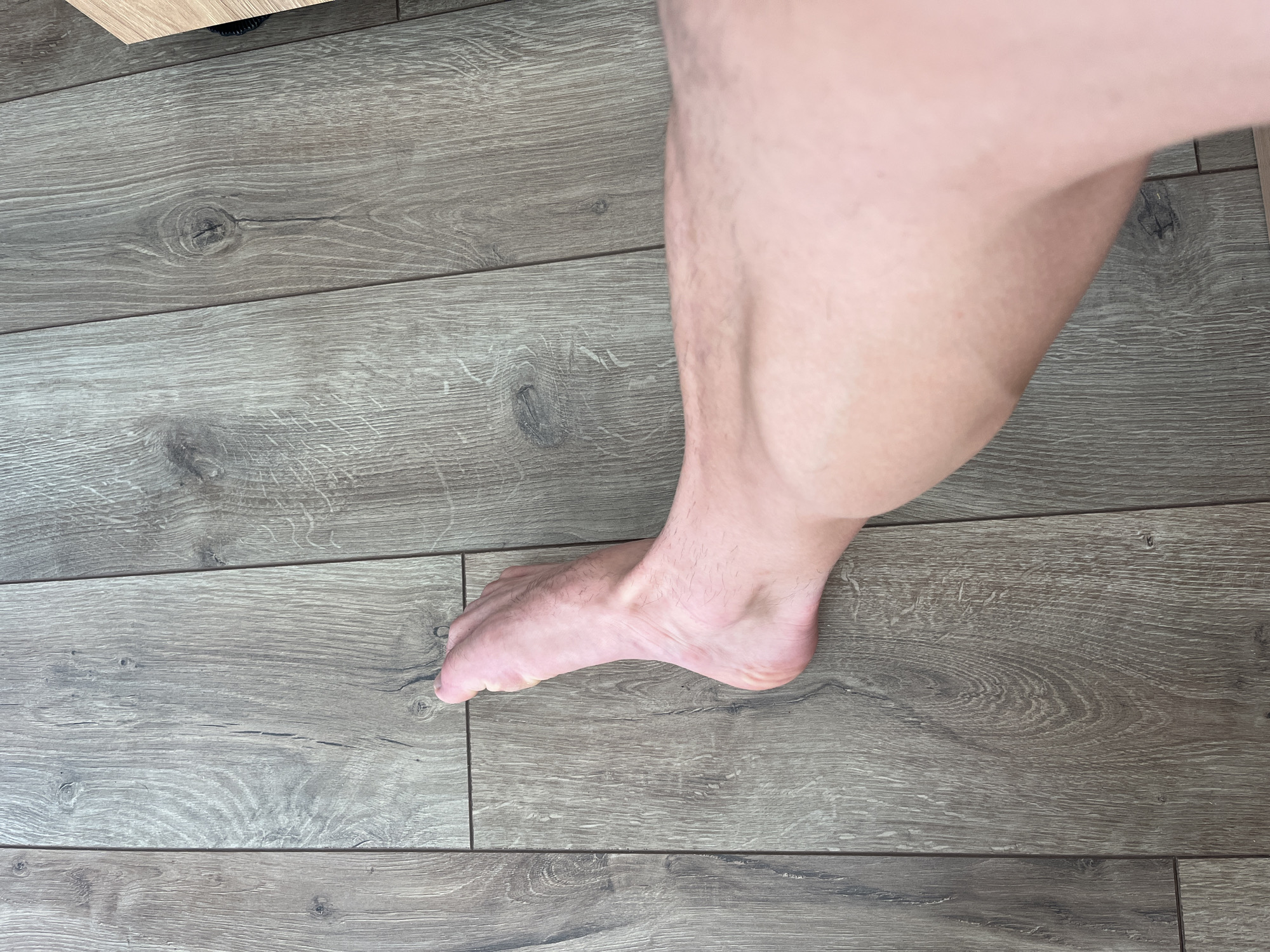 A close-up, overhead shot shows a bare leg and foot positioned on a weathered, gray wood-plank floor. The leg extends from the bottom right of the frame towards the left, where the foot is visible. The foot is pointed slightly upward and to the left. The wood planks are laid horizontally in the image and show a distinct grain pattern with knots and cracks. The lighting is consistent throughout the scene, suggesting an indoor environment. The overall impression is one of domestic normalcy.
