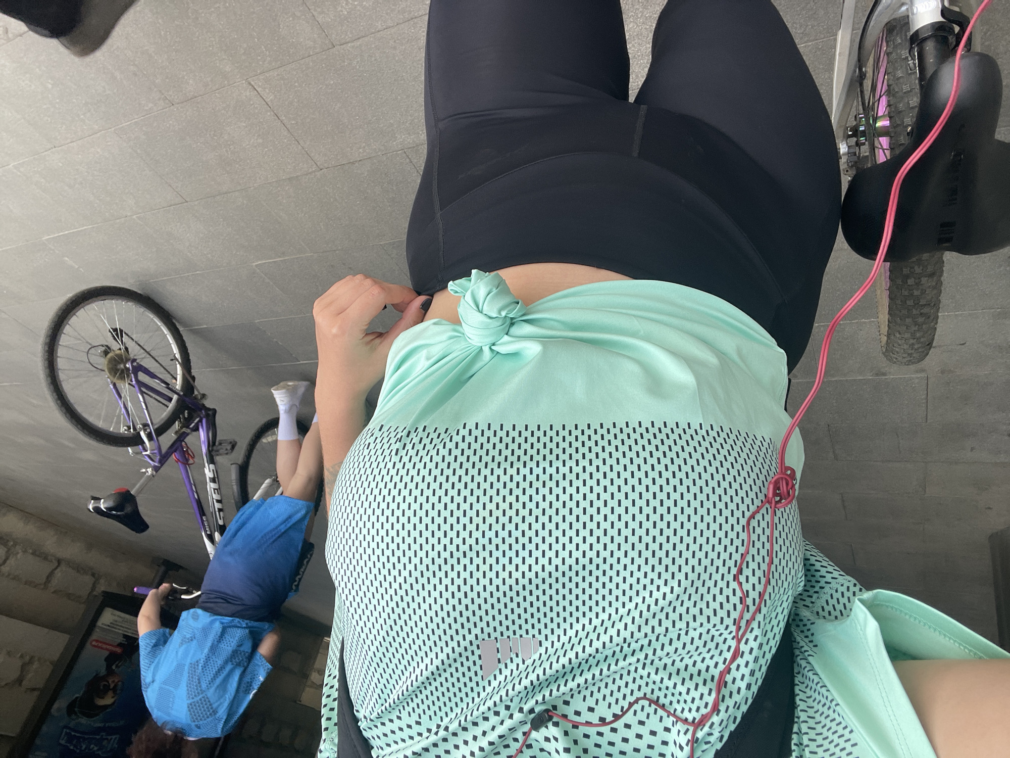 A street scene in Moscow, Russia, presents a partially visible person wearing black leggings and a light green t-shirt with a tied front. They are positioned near a bicycle with pink wiring. In the background, a child in blue shorts and a blue shirt lies next to another bicycle, seemingly on a sidewalk. The environment consists of a stone-tiled surface and a vertical wall. A poster with a graphic is also visible near the child.