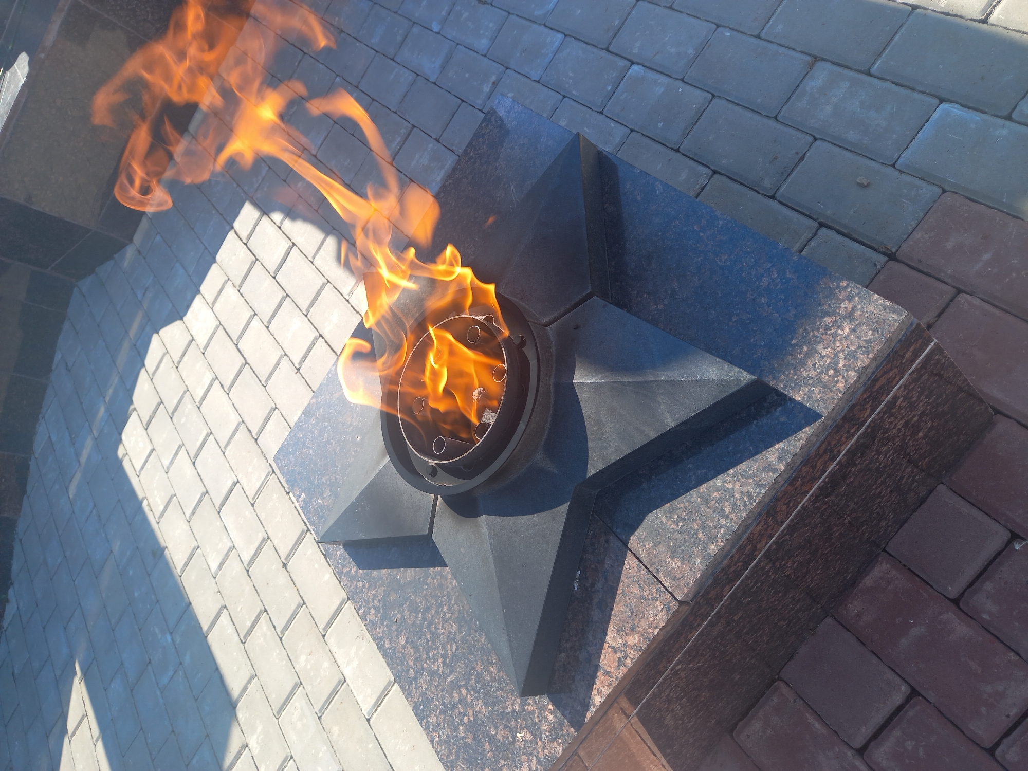 A close-up, high-angle view shows an eternal flame in a memorial setting. The flame burns brightly in a dark metal receptacle, which is set atop a black, granite-like star-shaped monument. The monument is surrounded by paving stones, some light grey and others a reddish-brown. Shadows from an unseen object stretch across the grey paving stones. In the background, darker stone structures, possibly part of a larger monument or building, are visible. The scene suggests a solemn and commemorative location, likely in a public space within the city of Sterlibashevo, Russia.