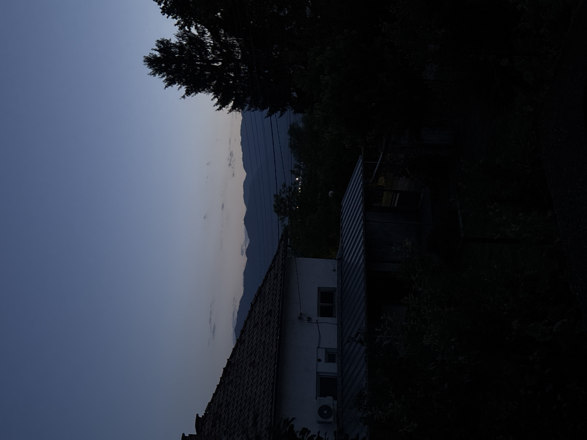 The image presents a serene, low-light landscape scene captured at dusk or twilight in Garmen, Bulgaria. The sky, a deep blue-grey overhead, gradually lightens towards the horizon, where distant mountains are silhouetted against the fading light. A faint, pinprick of light, possibly a star or planet, is visible in the upper sky.
Dense, dark foliage and tree branches frame the scene, occupying much of the foreground and the right side of the image. On the left, a light-colored house with a distinctive tiled roof is visible, featuring at least two windows and an outdoor air conditioning unit on its wall. Adjacent to it on the right is another structure, possibly a shed, clad in corrugated metal siding. Several power lines or cables stretch horizontally across the mid-ground, appearing above the distant mountain range.
Beyond the immediate structures, the mountainous terrain dominates the background, with the peaks and ridges forming a dark, undulating silhouette. Scattered artificial lights twinkle subtly in the distance, indicating a hidden settlement or village among the hills. The overall impression is one of quietude and the peaceful transition from day to night in a rural setting. No specific activity or individuals are depicted. A very faint, unreadable sign is present on the corrugated metal structure.