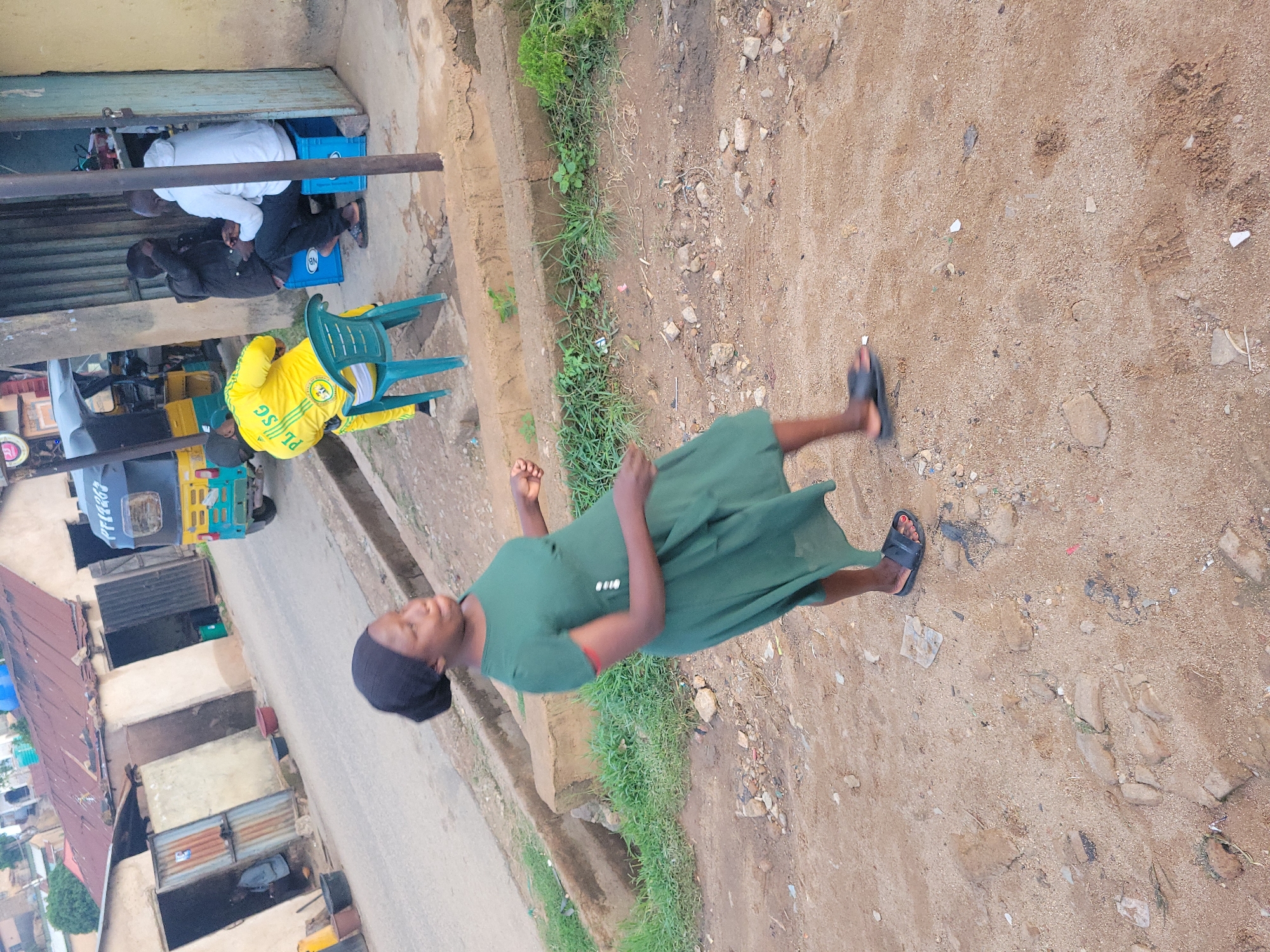 This image captures a street scene in Jos, Nigeria.
In the foreground, a dark-skinned woman with a dark head covering and a dark green, short-sleeved, knee-length dress walks toward the right on an unpaved, sandy-brown surface. This surface is uneven and contains scattered small rocks, dirt, and minor debris. She is wearing dark-colored slides on her feet, and red nail polish is visible on her left foot's toes. Her head is turned slightly to the left, and she displays a smile. Her right arm is slightly raised.
Behind the woman, a paved road extends diagonally from the lower left to the upper right. A concrete gutter or drainage channel runs parallel to the road, containing some green foliage.
Along the right side of the road, three men are seated near a building with a partially open, turquoise-colored corrugated metal door. One man wears a yellow tracksuit with green stripes and the inscription "PL | SG" on the back; he is seated on a green plastic chair, facing away from the camera. The other two men are seated on blue plastic crates facing each other; one wears a black jacket, and the other a white hoodie. They appear to be engaged in conversation or viewing a device. A yellow and green auto-rickshaw (Keke Napep) is partially visible on the road behind these men, with some unreadable white text on its rear top section.
To the left of the road, a row of modest, single-story buildings with corrugated metal roofs and various types of doors are visible. Adjacent to these buildings are several containers, including black buckets, a yellow bucket, and a red basin. In the background, more buildings and trees are present, suggesting a populated urban or semi-urban environment under an overcast sky.
