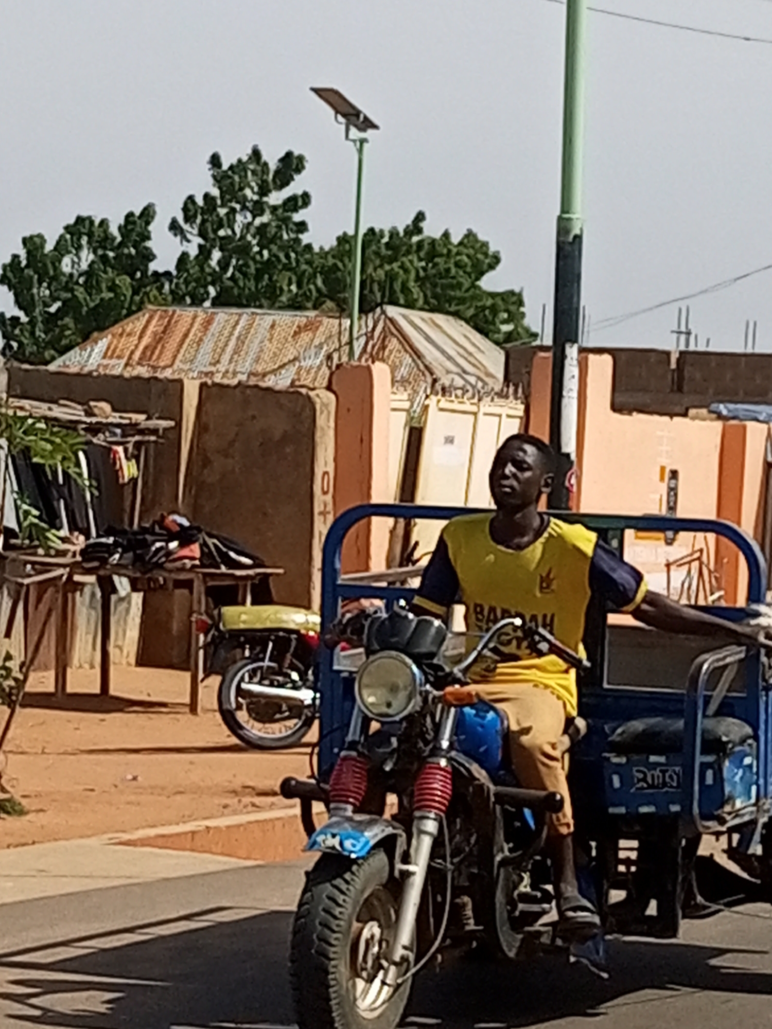 A man is riding a blue and black three-wheeled motorcycle in Tempure, Nigeria. He is wearing a yellow and blue t-shirt with the word "BARRAH" printed on it. He is also wearing tan pants. The motorcycle has a blue seat and a blue frame. The man is looking to his left. In the background, there are some buildings and trees. There is also a solar-powered street lamp. The scene is outdoors during the day.