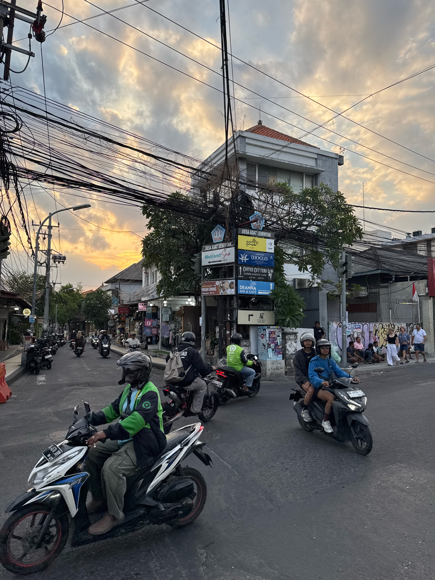 The image depicts a street scene in Kuta, Indonesia, with a focus on a road filled with motorbikes and several pedestrians. The sky, visible at the top of the frame, is overcast with golden light, suggesting either sunrise or sunset. A complex network of overhead electrical wires dominates the upper portion of the image, strung between utility poles and buildings.
In the foreground, a man wearing a green jacket with a GOJEK logo is riding a white and blue Honda motorbike. He is wearing a helmet and khaki pants. Behind him, another motorbike carries two individuals, both wearing helmets, with the rider in a blue shirt and the passenger in a darker shirt. Further down the street, numerous other motorbikes are visible, some parked and others in motion.
On the right side of the street, a group of people are sitting and standing near a building with graffiti on its lower section. A few individuals are walking along the sidewalk. The buildings lining the street appear to be commercial establishments, with signs indicating businesses such as "DESA ADAT SEMINYAK," "WARUNG DJOGLO," and "COFFEE CUTTERS." The overall atmosphere suggests a busy urban environment.