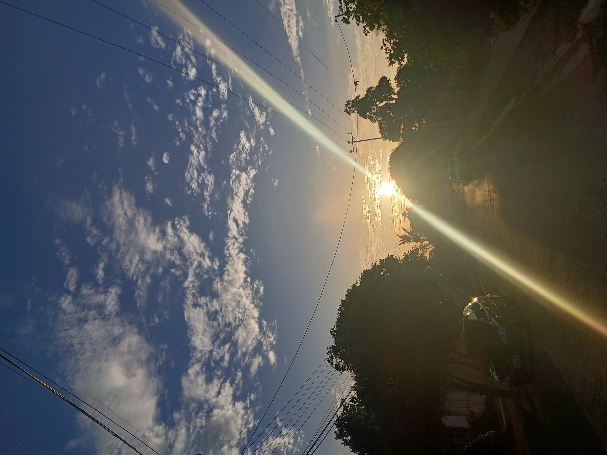 The image shows a street scene in Juan Griego, Venezuela, at sunset. The view is oriented vertically, with the sky occupying the left portion of the frame. Altocumulus clouds are scattered across a blue sky. Power lines stretch across the sky. The sun is setting over the road, casting a bright glare across the image. There are trees lining the right side of the road. A vehicle is parked on the right.