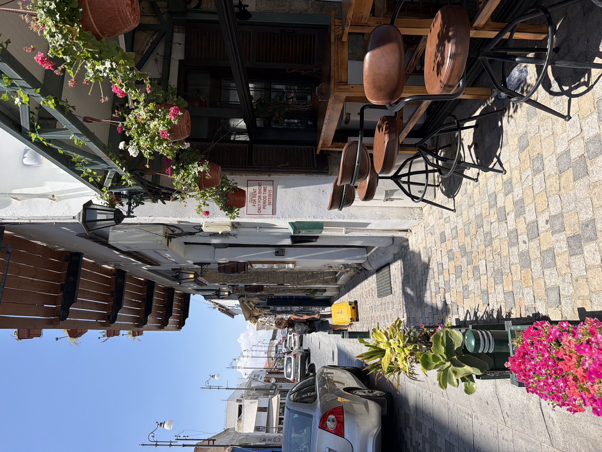 In an outdoor urban setting on a sunny day, a woman with dark curly hair, wearing a dark tank top and shorts, stands on a cobblestone street pulling a bright yellow rolling suitcase. She is facing away from the camera and towards the right side of the frame. The street is lined with buildings that have awnings and hanging flower baskets. A silver sedan is parked on the left side of the street. In the foreground, a row of planters contains green plants and vibrant pink flowers. A wooden table with three brown leather bar stools is positioned outside a building on the right. A red sign is visible on the white wall of the building, stating "ROOMS FOR RENT ONLY FOR SHORT PERIOD TIME 99753915." The sky is clear and blue.
