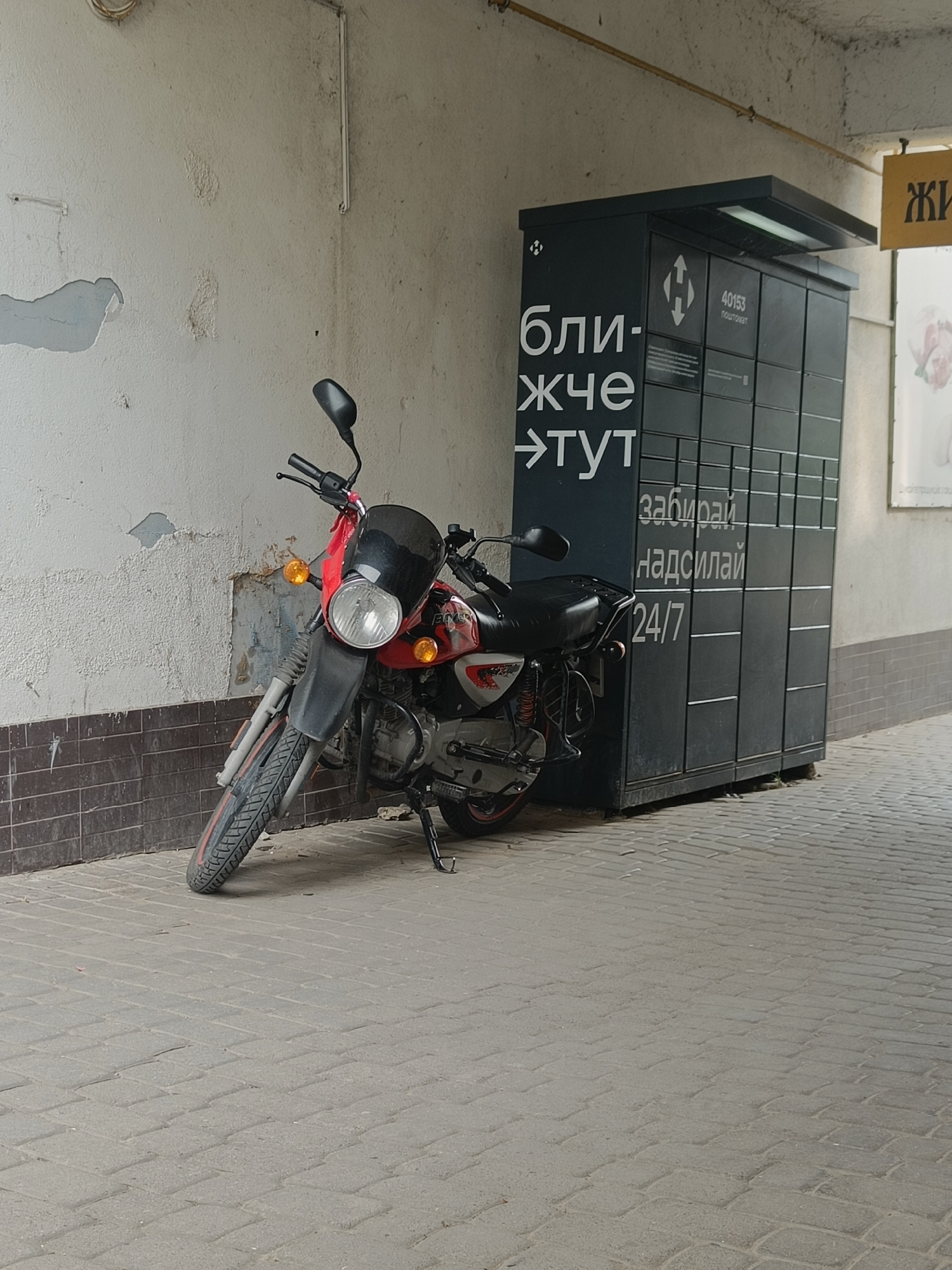 A red and black motorcycle is parked on a brick sidewalk in front of a black parcel locker unit. The parcel locker unit has white text that reads "бли-жче ->тут", "забирай", "надсилай", and "24/7". Above the text, there is a label that reads "40153 ПОШТОМАТ". The motorcycle has a black windscreen and a black seat. The wall behind the motorcycle is textured and painted white, with some areas of peeling paint. To the right of the parcel locker, there is a yellow sign with black text that reads "ЖИ". The environment appears to be an outdoor urban setting.