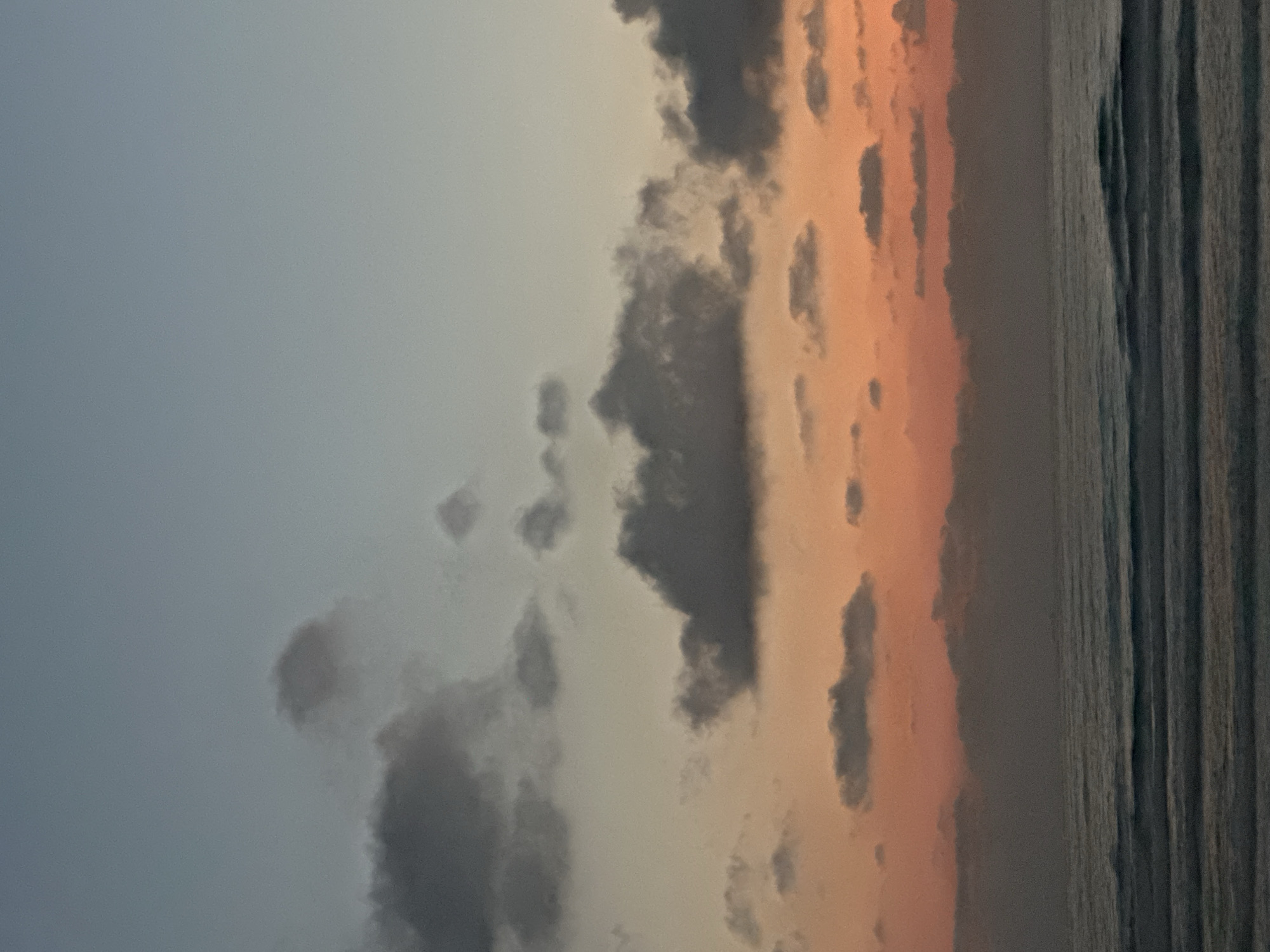 This is a landscape photograph taken in Haifa, Israel, likely at dusk. The image depicts a serene coastal scene with a focus on the sky and the sea. The sky is divided into two distinct sections: the left side is a pale blue, transitioning to a soft, peachy orange hue on the right, indicating the setting sun. Dark, wispy clouds are scattered across the sky, adding depth and texture to the celestial display.
The sea occupies the right portion of the frame, characterized by a dark, rippling surface that reflects the colors of the sky. The texture of the water suggests a gentle breeze or calm conditions. The horizon line is not clearly defined, as the sea and sky blend together, creating a sense of vastness.
There are no people or man-made objects visible in the frame, emphasizing the natural beauty of the location. The overall mood is peaceful and contemplative, evoking a sense of tranquility often associated with sunsets. The color palette, dominated by blues, grays, and warm oranges, contributes to the atmospheric quality of the image. The photograph captures a moment of natural beauty, highlighting the interplay of light and color in a coastal environment.