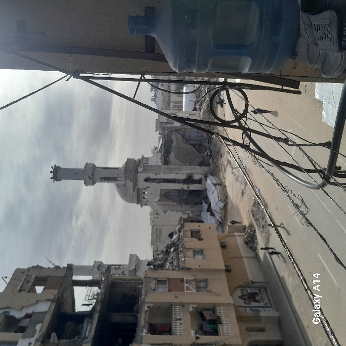 The image shows a destroyed urban landscape, with buildings reduced to rubble and a partially standing mosque in the background. Electrical wires crisscross the scene, and a blue water bottle and other objects are visible in the foreground, suggesting the photo was taken from a higher vantage point, like a balcony. The devastation is extensive, implying a scene of conflict or natural disaster.