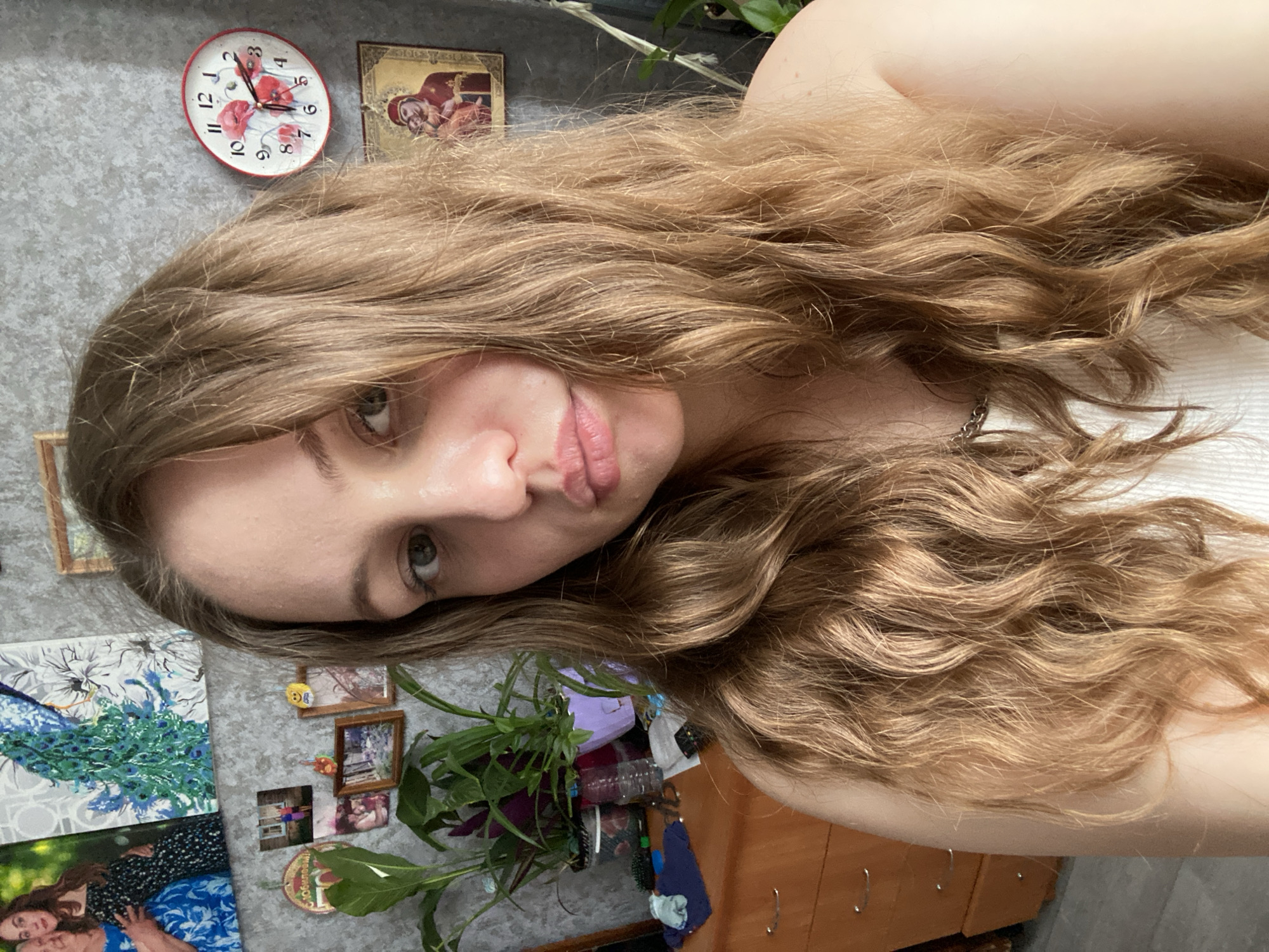 A young adult female is depicted from the chest up, positioned centrally and looking directly at the camera with a slight pout and green eyes. She has long, wavy, light brown hair that cascades over her shoulders and chest. She is wearing a white ribbed tank top, with her bare shoulders visible. A silver chain necklace is around her neck.
The scene is an interior setting located in Марковский, Russia. The background consists of a textured grey wall, densely decorated. On the left, two large framed items are visible: one is a photograph of two women embracing, and the other is a vibrant artwork featuring a blue and green peacock feather design against a white floral pattern. Below these, a cluster of smaller framed photographs shows people in various settings, including two individuals outdoors and a group indoors. A golden circular plaque bearing Cyrillic text, including "50 ЛЕТ" (50 Years), and small decorative magnets are also on the wall.
To the right, a circular wall clock with red poppy illustrations displays approximately 11:15. Directly below the clock, a framed religious icon depicting Madonna and Child figures is affixed to the wall. Further to the right, a tall, slender plant is partially visible.
In the lower left portion of the image, a light brown wooden dresser with silver handles is visible. The top surface of the dresser holds several potted plants in dark and purple containers. Other items on the dresser include a clear plastic water bottle, hairbrushes, a dark blue fabric item, and a small white object.