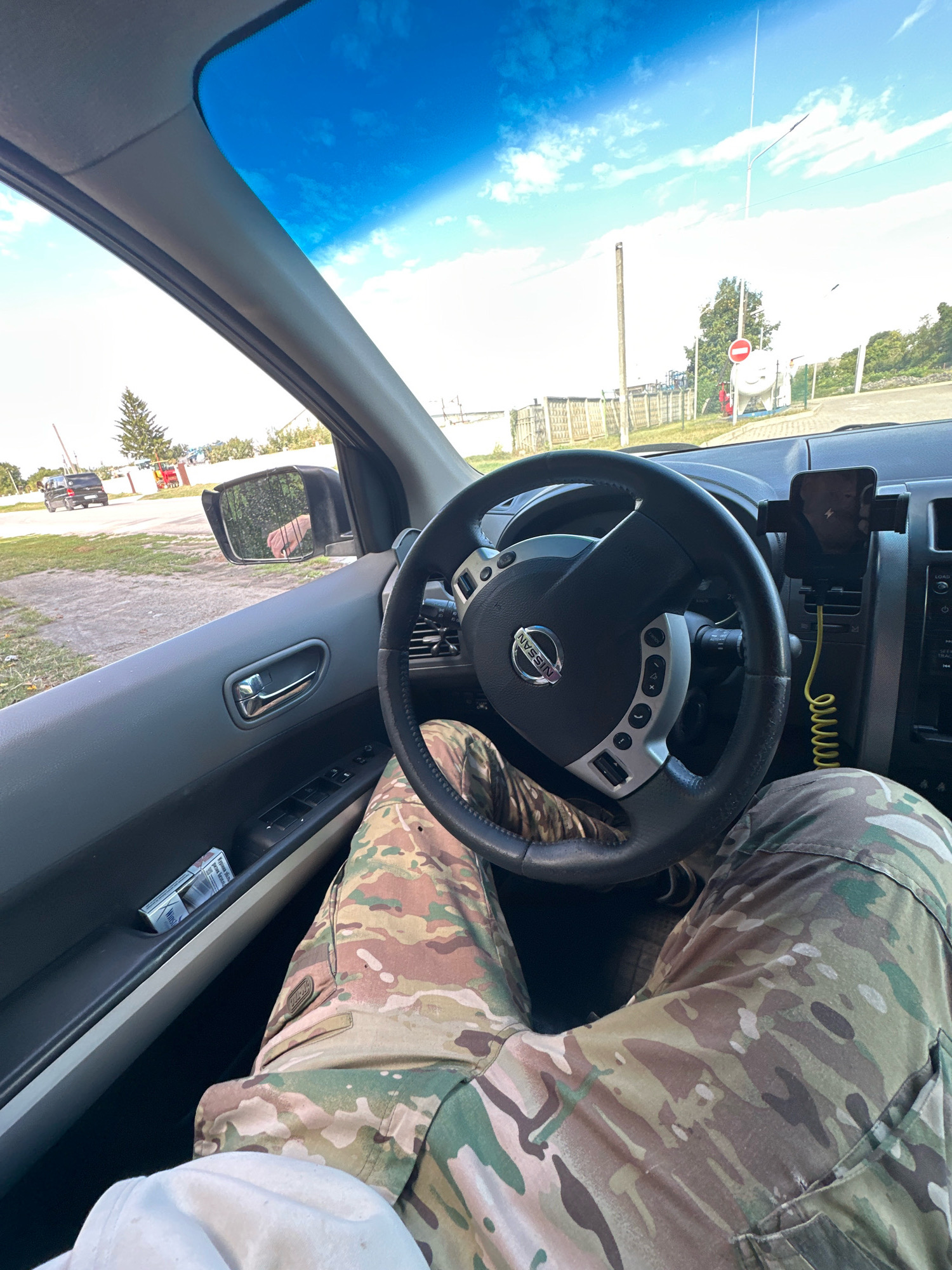 The image shows the interior of a car, looking out through the windshield and side window. The driver's legs, clad in camouflage pants, are visible in the foreground. The steering wheel with the Nissan logo is prominent. A smartphone is mounted on the dashboard and is connected to a charger. A pack of cigarettes is visible in the door pocket. Outside the car, a rural landscape with trees, a fence, and a gas storage tank can be seen under a blue sky with scattered clouds. The location is identified as Shatava, Ukraine.