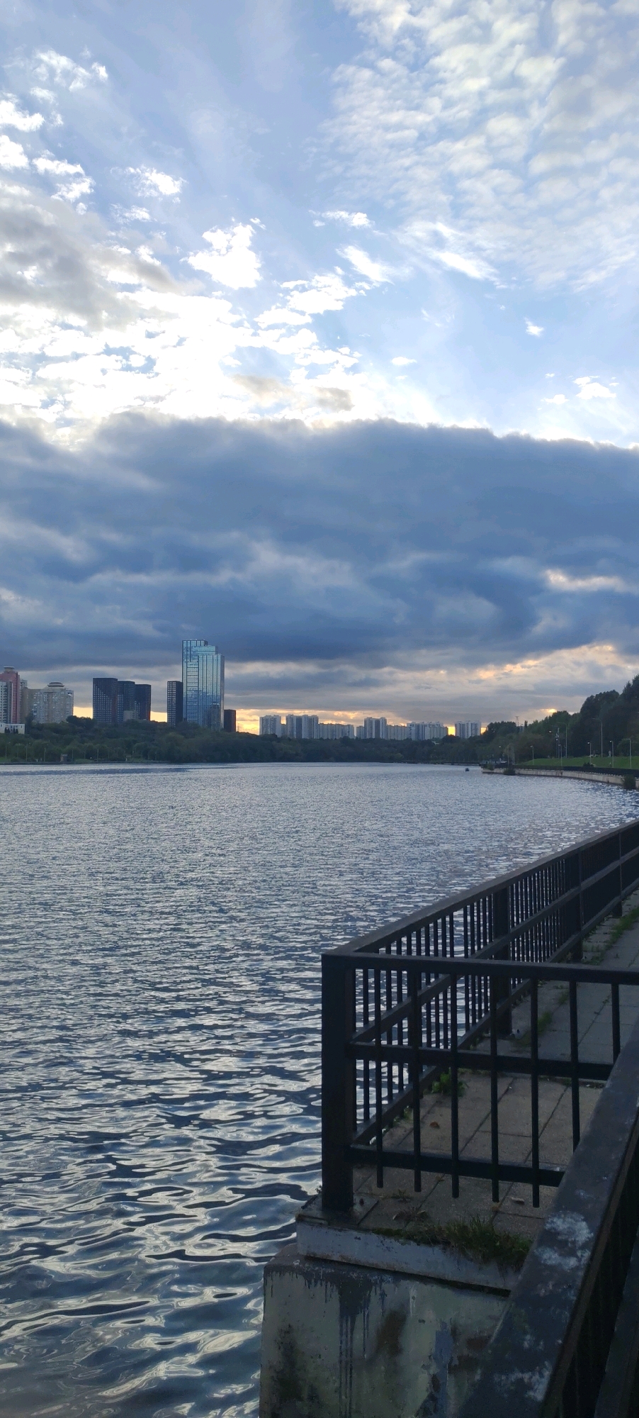 The image depicts a wide view of a body of water, likely a river or lake, with a city skyline in the background. The water is rippled, reflecting the cloudy sky. In the foreground, a dark metal railing runs along the edge of a concrete embankment, creating a sense of depth and perspective. The railing is partly obstructed by concrete supports.
The city skyline features a mix of modern high-rise buildings and apartment blocks, interspersed with trees. One prominent building is a sleek, glass-covered skyscraper. The sky above is a dramatic expanse of clouds, with patches of lighter sky suggesting either sunrise or sunset. The overall mood is serene and perhaps a bit overcast.
This scene is consistent with a view from the Strogino floodplain or embankment in Moscow, Russia. The distinctive architecture of the buildings, the presence of a large body of water, and the park-like setting with pathways and greenery are characteristic of this area. The time of day appears to be either early morning or late evening, given the lighting conditions and the cloud formations. There are no people visible in the image.