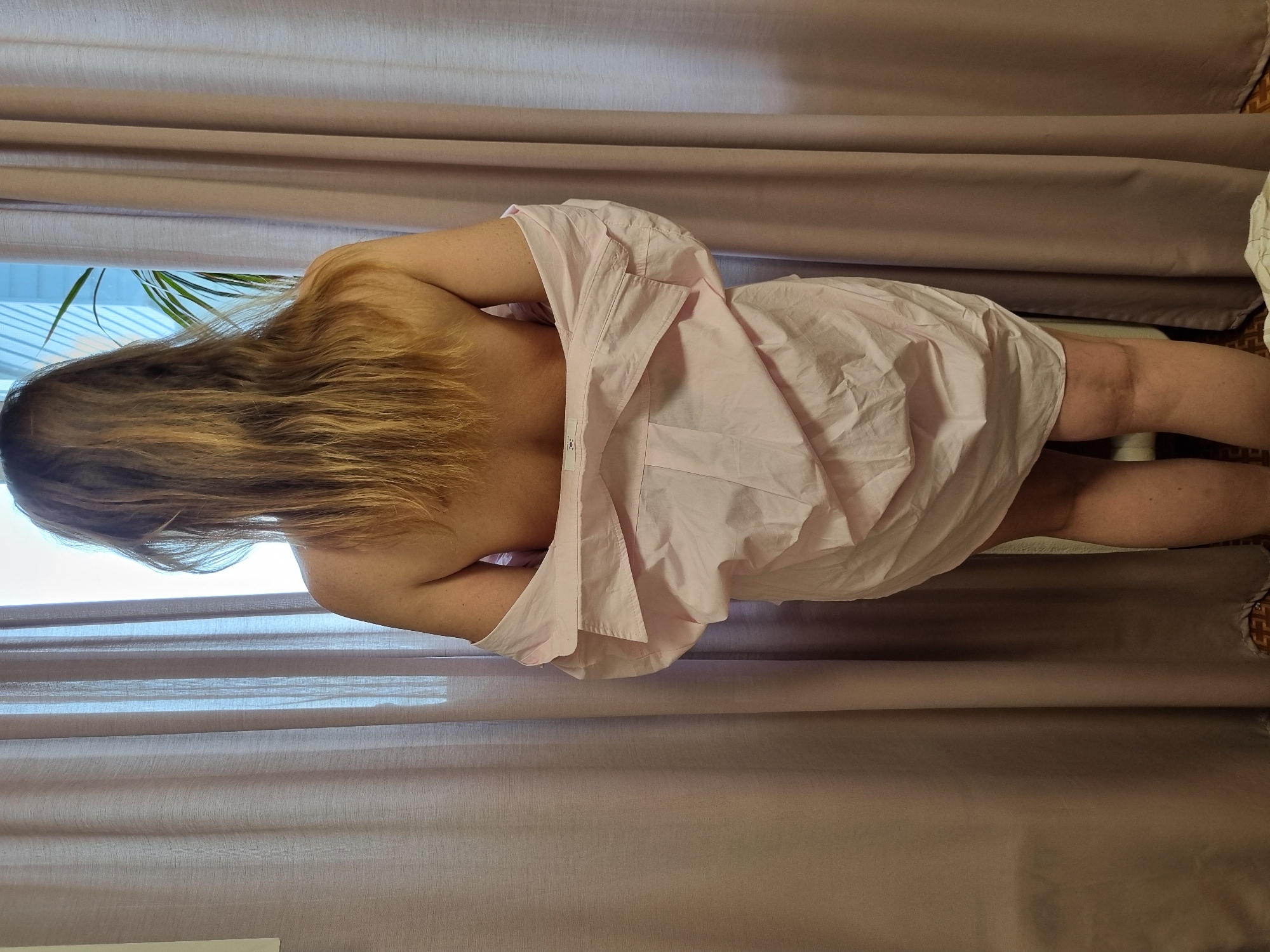 A woman with long blonde hair is standing in front of a window with closed beige curtains. She is wearing a light pink button-down shirt that partially covers her body. The window is partially open, revealing a glimpse of the outside, possibly a residential area, as suggested by the presence of a plant. The scene appears to be indoors, possibly in Chaikovsky, Russia.