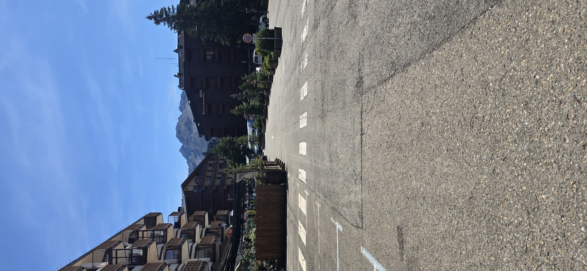 The image captures a street scene in Sallent de Gállego, Spain, under a clear blue sky with distant mountains. The foreground features a paved road with a pedestrian crossing marked by white stripes. To the left, a multi-story building with numerous balconies is visible, adorned with dark brown railings. Adjacent to this building is a wooden fence enclosing an outdoor seating area, likely a restaurant or cafe, with greenery and tables. Across the street, more multi-story buildings with dark facades and balconies are present, along with several evergreen trees. Parked vehicles, including a white van and a car, are visible on the right side of the road. A circular sign with red and blue symbols indicating a restriction is also situated on the right. The overall scene depicts a tranquil, daytime setting in a mountainous town.