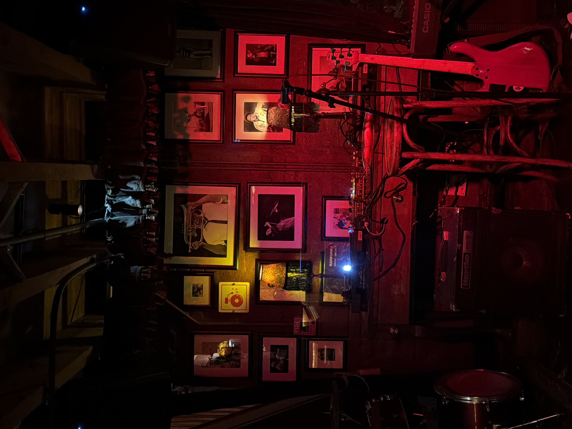 The image depicts a dimly lit, cluttered interior. A wall is adorned with framed photographs and other memorabilia. A red electric guitar rests on a shelf near a "CASIO" keyboard. Various pieces of musical equipment are visible, including a microphone, wires, an amplifier, and a drum. To the left is a wooden staircase leading to an unseen upper level. A spotlight shines on the wall. A poster for "MANKUNKU TADHAI" is also visible. It's hard to say if the picture was taken in Cape Town, as the location is not evident.