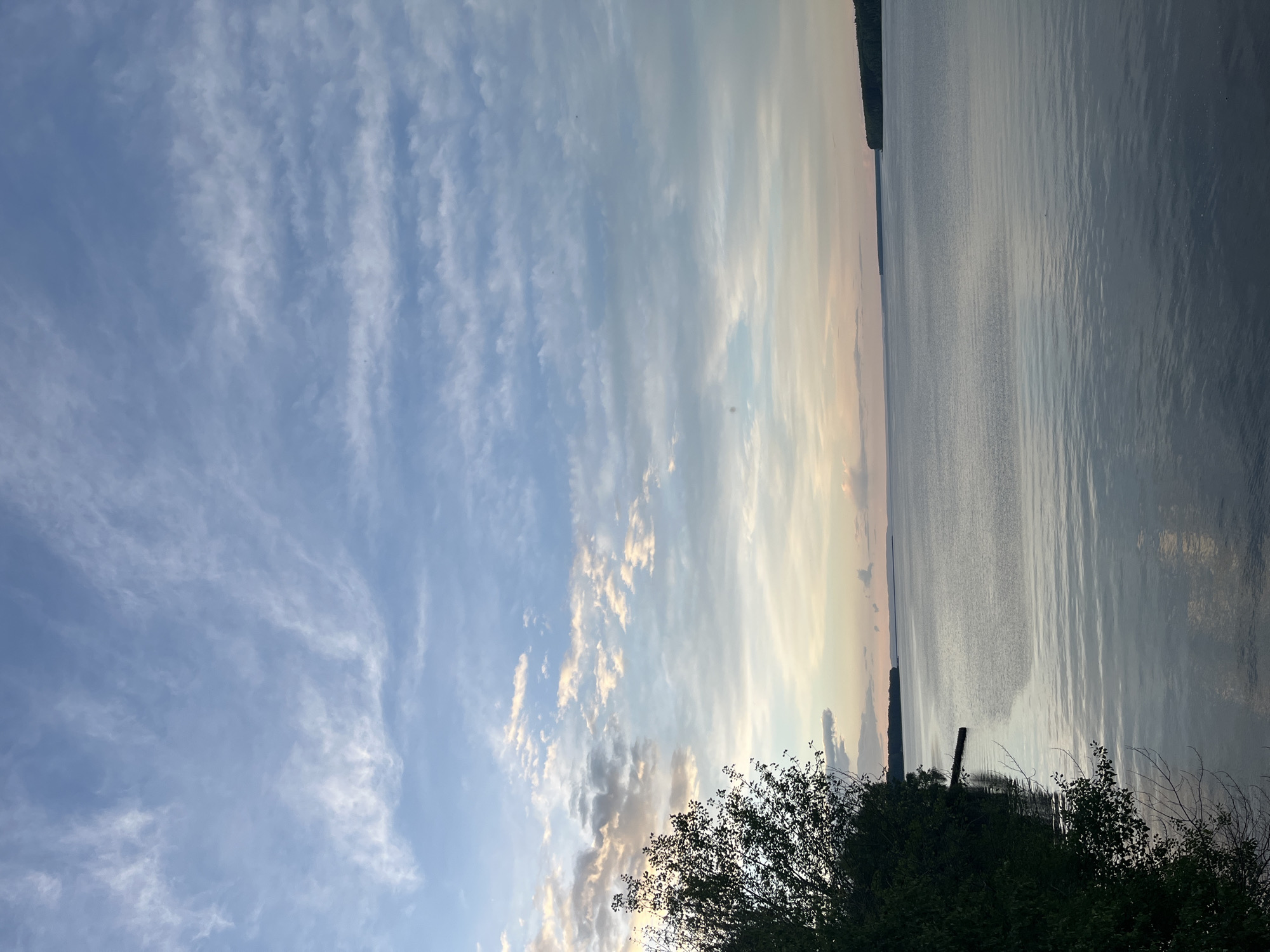 The image captures a waterfront scene in Chaikovsky, Russia, during twilight. The sky displays cirrus clouds, transitioning from blue to a light orange hue on the horizon. A body of water, likely the Kama River, stretches horizontally across the frame, reflecting the colors of the sky. On the right, the opposite shore is visible as a dark silhouette of trees and land. In the foreground, bushes with dark foliage are present. A single wooden post is partially visible along the waterline. The scene has a tranquil and reflective atmosphere.