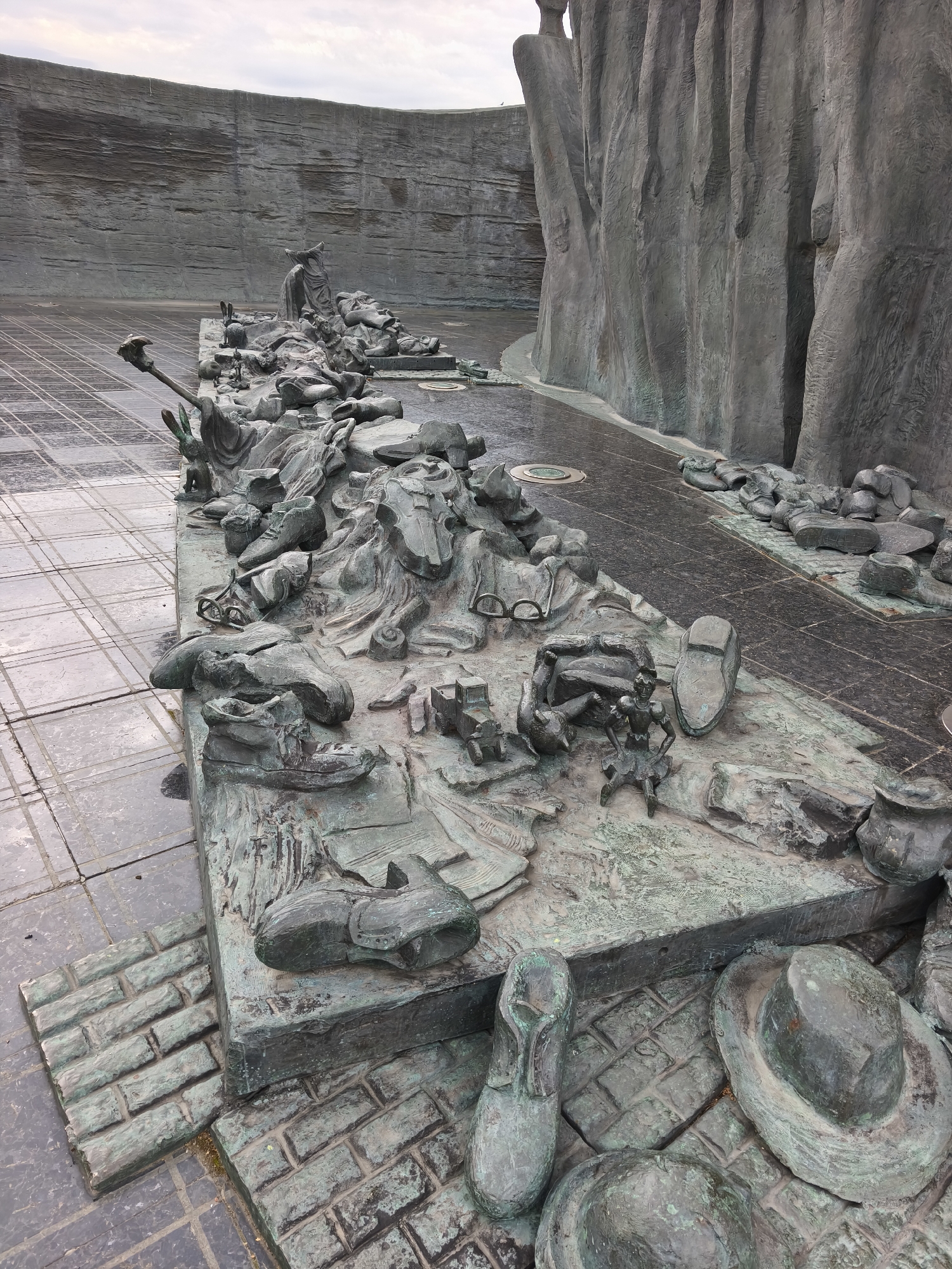The image depicts a large, abstract bronze sculpture located in Moscow, Russia. The sculpture is situated in an open, paved area with a large, textured wall forming the backdrop. The ground is a dark, patterned surface, possibly stone or concrete tiles, with some areas appearing wet, suggesting recent rain.
The sculpture itself is a dense, chaotic arrangement of various human figures, clothing, and everyday objects rendered in a stylized, somewhat grotesque manner. Scattered throughout the composition are numerous shoes, hats, eyeglasses, and what appear to be parts of figures in various states of distress or disarray. The bronze has a verdigris patina, indicative of age and exposure to the elements.
In the foreground, there is a collection of shoes and hats, including a large fedora. A single shoe is prominently displayed on a raised platform made to resemble brickwork. To the left, the paved ground transitions to a simulated brick walkway.
Further into the sculpture, various fragmented figures are visible, some appearing to be lying down, others in more dynamic poses. Among the scattered items, there are objects that could be interpreted as furniture or tools. The overall impression is one of a tragic event, with personal belongings scattered and figures appearing to be overwhelmed or lost.
The scale of the sculpture is substantial, dominating the visible area of the paved plaza. The rough, textured wall behind the sculpture creates a sense of enclosure and adds to the somber atmosphere. The sky above is overcast, contributing to the muted color palette of the scene.