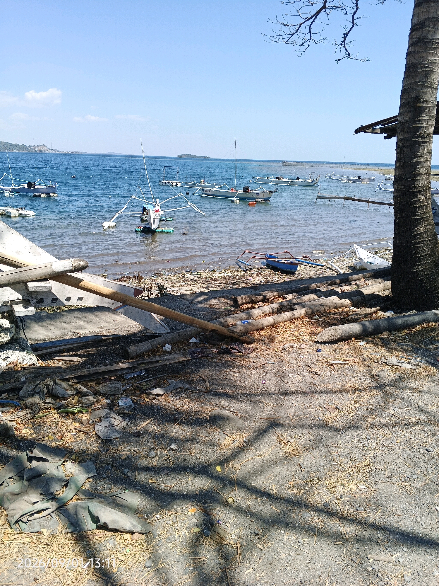 The image shows a beach scene in Kupa, Indonesia, on a clear, sunny day. Several small, traditional outrigger boats are anchored in the calm blue water. The shoreline is littered with debris, including plastic waste, dried leaves, and logs, suggesting a lack of regular cleanup. In the foreground, a large palm tree trunk is visible on the right, and shadows from its fronds cast patterns on the sandy ground. Some dilapidated structures and discarded materials are also present on the beach. The overall impression is of a somewhat neglected coastal area.