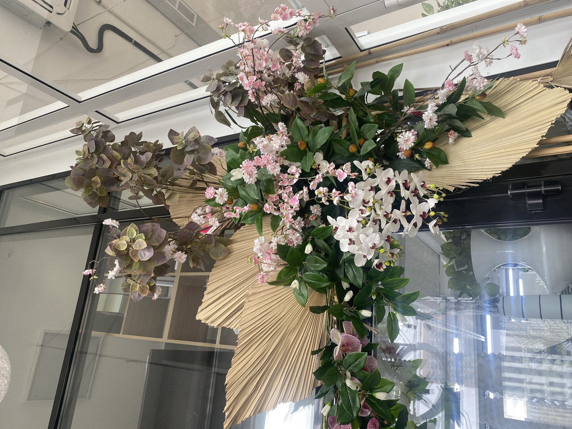 A floral arrangement is mounted on a glass wall in what appears to be an indoor location. The arrangement consists of pink and white flowers, green foliage, and large dried palm leaves. The flowers and foliage are arranged in a cascade, with the palm leaves providing a backdrop. Behind the glass wall are glimpses of interior spaces with shelves and other decorative items. Based on the context provided, this location is possibly in the city of Yekaterinburg, Russia.