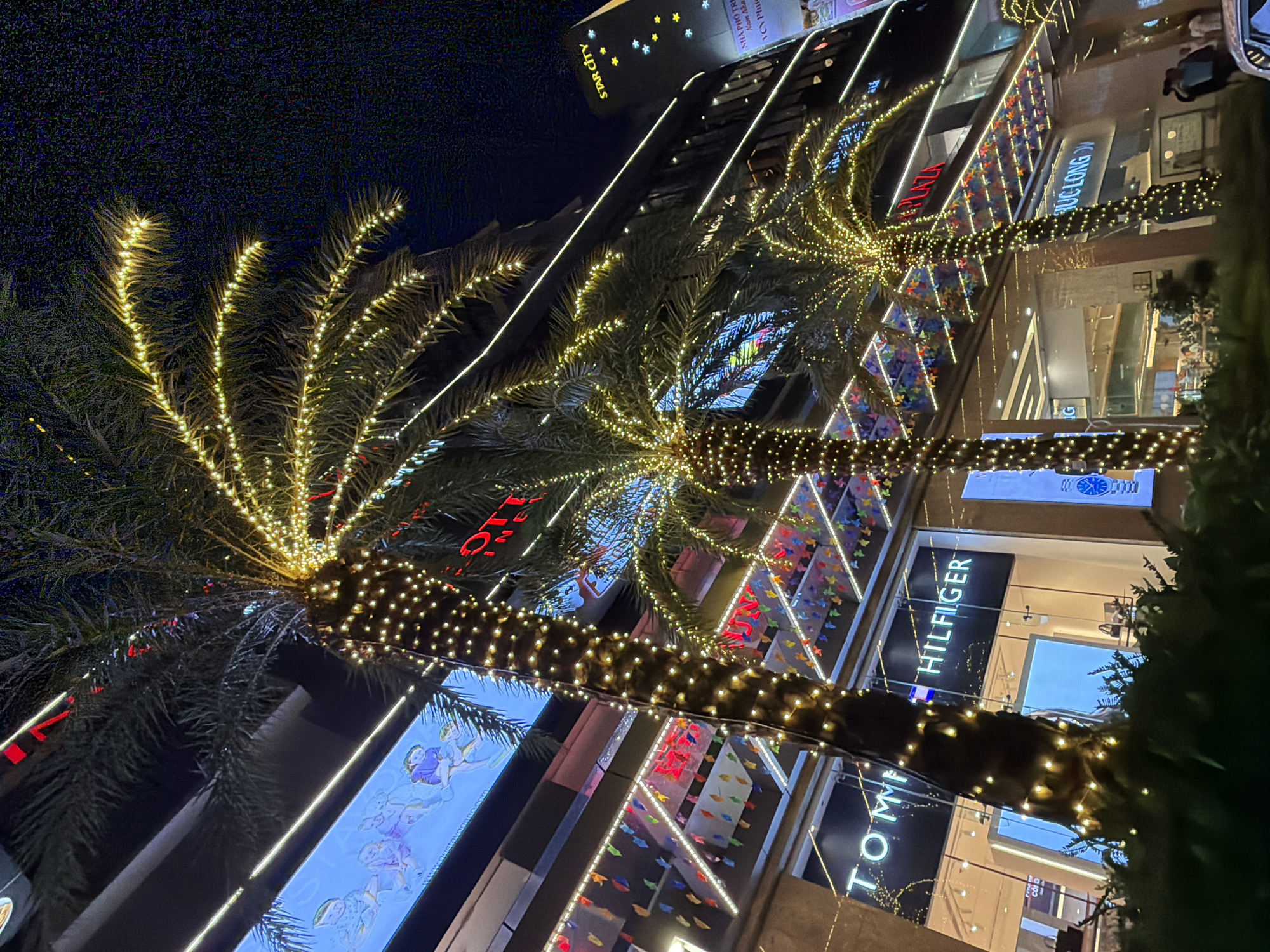 The image depicts a nighttime scene in Nha Trang, Vietnam, focusing on a section of a modern building facade adorned with Christmas lights. Three palm trees, wrapped in numerous small, warm-toned lights, are prominent in the foreground, their fronds extending upwards. The building behind them features large illuminated signs, including "TOMMY HILFIGER" and "STAR CITY". Above the Tommy Hilfiger sign, a digital display shows animated figures. Various smaller, colorful decorative flags are strung across the building's upper levels. The overall impression is of a festive, urban commercial area at night.