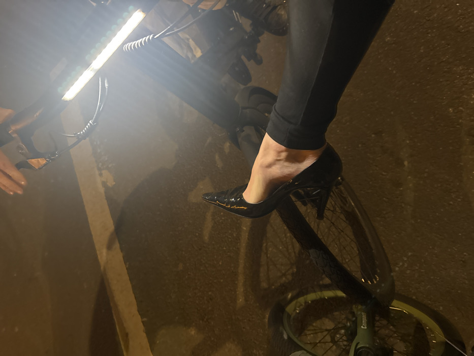 The image depicts a nighttime scene in the city of Paris, France, identifiable by the distinctive "Compact" branding on a light green component of a bicycle, consistent with a Vélib' Métropole bike, along with its yellow-green wheel rims. The scene is illuminated by artificial light, primarily from a bright, rectangular LED light attached to the front of the bicycle, which emits intense white light causing significant glare in the upper right quadrant.
A person's lower leg and foot are prominently featured, extending horizontally across the mid-section of the frame. The person is wearing black trousers or leggings and a black, shiny, pointed-toe high-heeled pump. The foot is partially dislodged from the shoe, revealing the heel and arch. This foot is positioned above the dark-colored frame of the bicycle. A hand, likely belonging to the same individual, is visible near the bicycle's handlebars. The ground appears to be dark, wet asphalt or pavement, reflecting the light and showing a faint white line in the foreground. Shadows from the bicycle and other unseen elements are cast upon the wet surface.