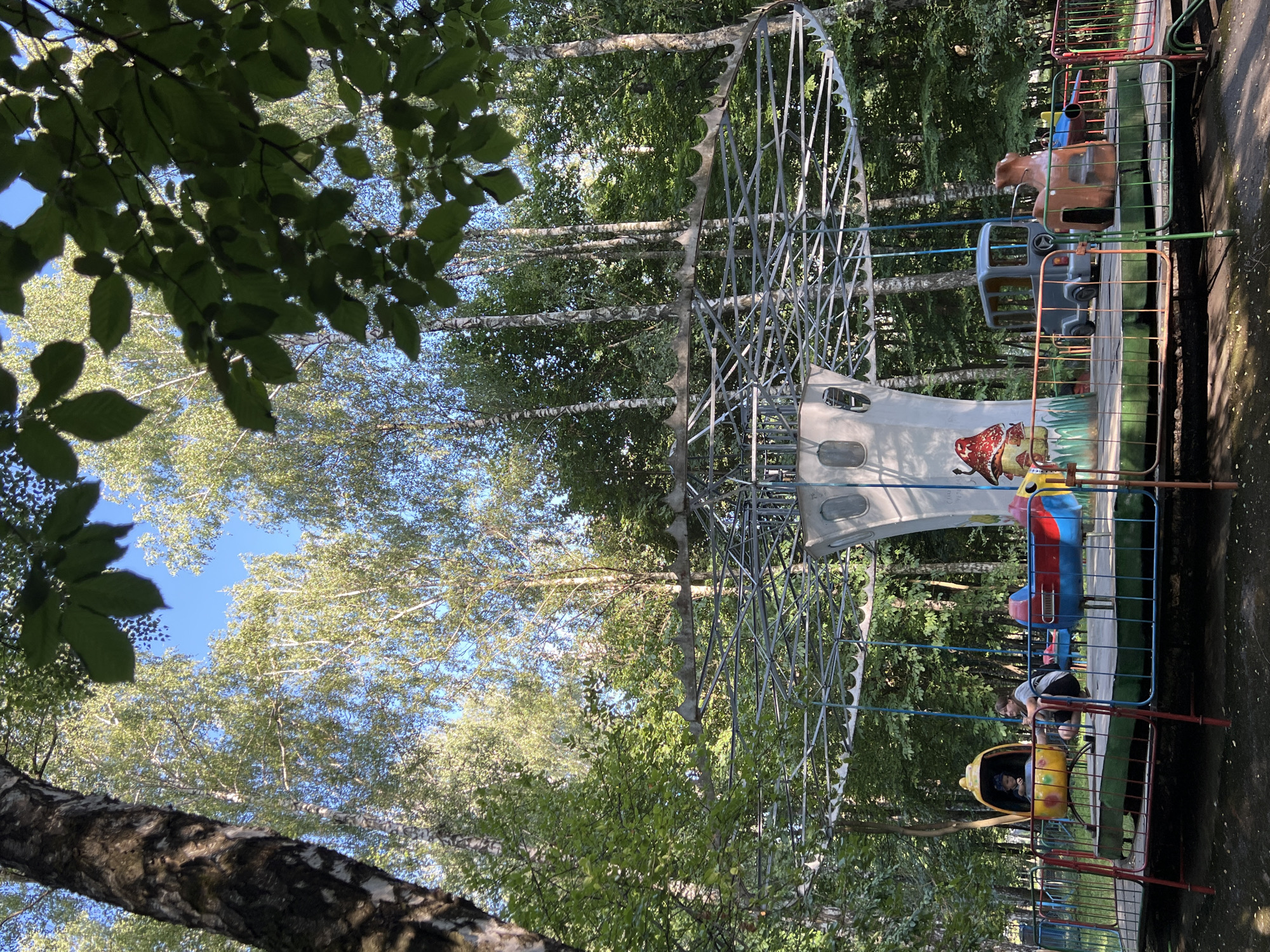 This image depicts an amusement park ride on a sunny day. The ride features several colorful, airplane-shaped gondolas suspended from a circular metal frame. In the foreground, two children are visible inside one of the gondolas, appearing to enjoy the ride. Another gondola is seen further back, occupied by at least one person. The ride is surrounded by lush green trees, suggesting an outdoor setting, possibly a park or a recreational area. The presence of the amusement ride and the clear sky indicate a pleasant day for outdoor activities. The scene is set in Ternopil, Ukraine. No text is visible in the image.