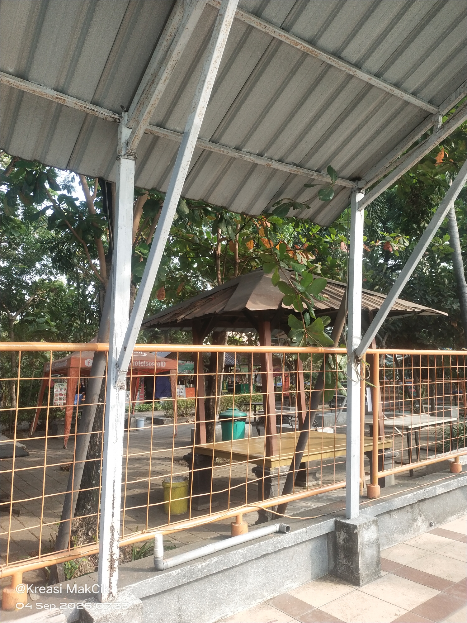 The image depicts an outdoor seating area with a corrugated metal roof supported by metal beams. A wooden gazebo with a dark roof is visible in the background, along with trees and foliage. An orange metal fence with square mesh encloses the area. Several tables and chairs are set up within the fenced area. The ground is paved with square tiles. In the foreground, a concrete structure with a pipe attached is present. The image is watermarked with "@Kreasi MakCik" and "04 Sep 2026 07:33", suggesting it was taken in Suko, Indonesia. There are no people visible in the image.