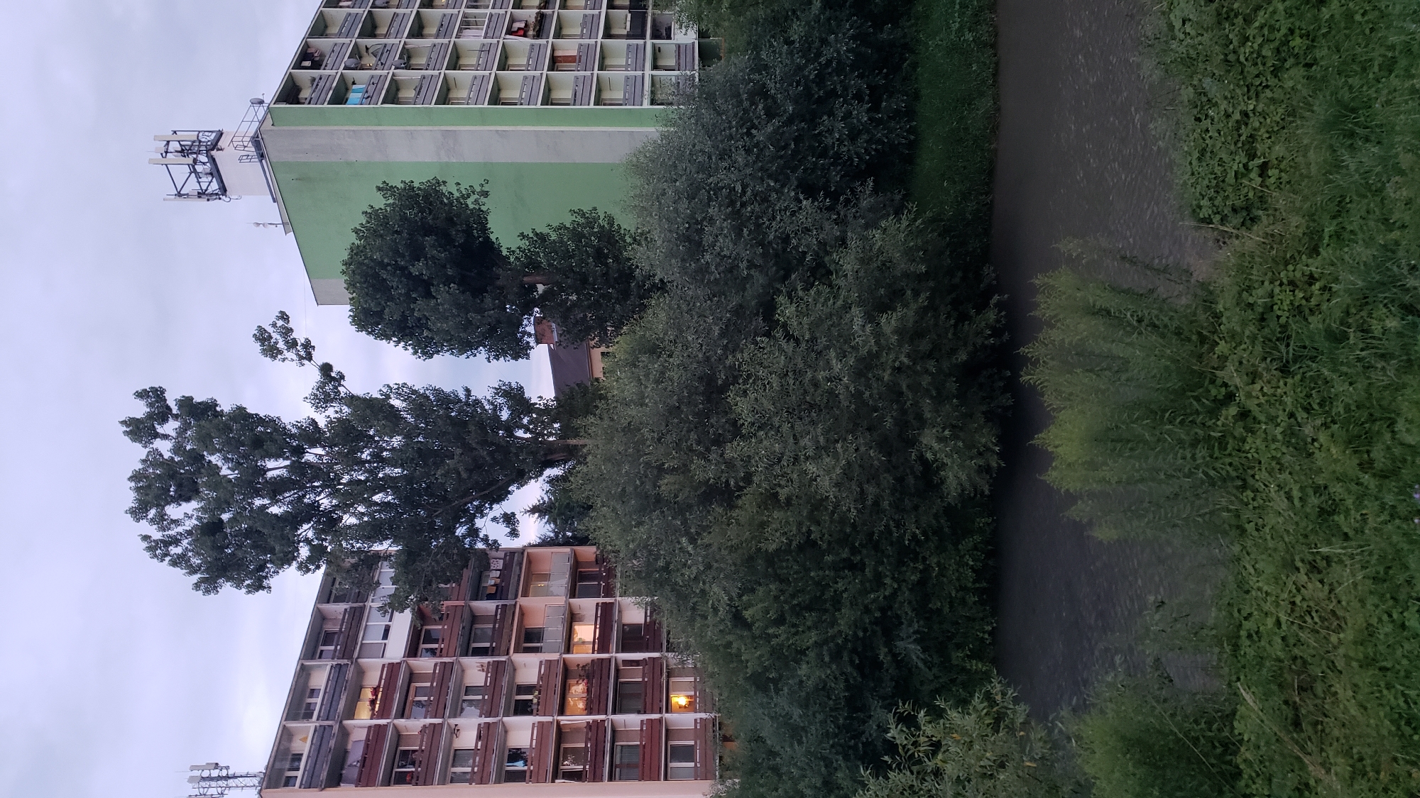 The image depicts a residential building with a light brown facade and balconies, situated next to a building with a green facade, both of which are multi-story apartment blocks. In front of the buildings and between them are dense trees and bushes. A body of dark water, likely a river, flows in the foreground, bordered by lush green vegetation. The sky is overcast and the scene appears to be in the evening or early morning. The buildings are typical of socialist-era architecture found in many Eastern European cities, including Prešov, Slovakia. There are no people or observable activities in the image. The overall impression is of an urban residential area with significant greenery.
