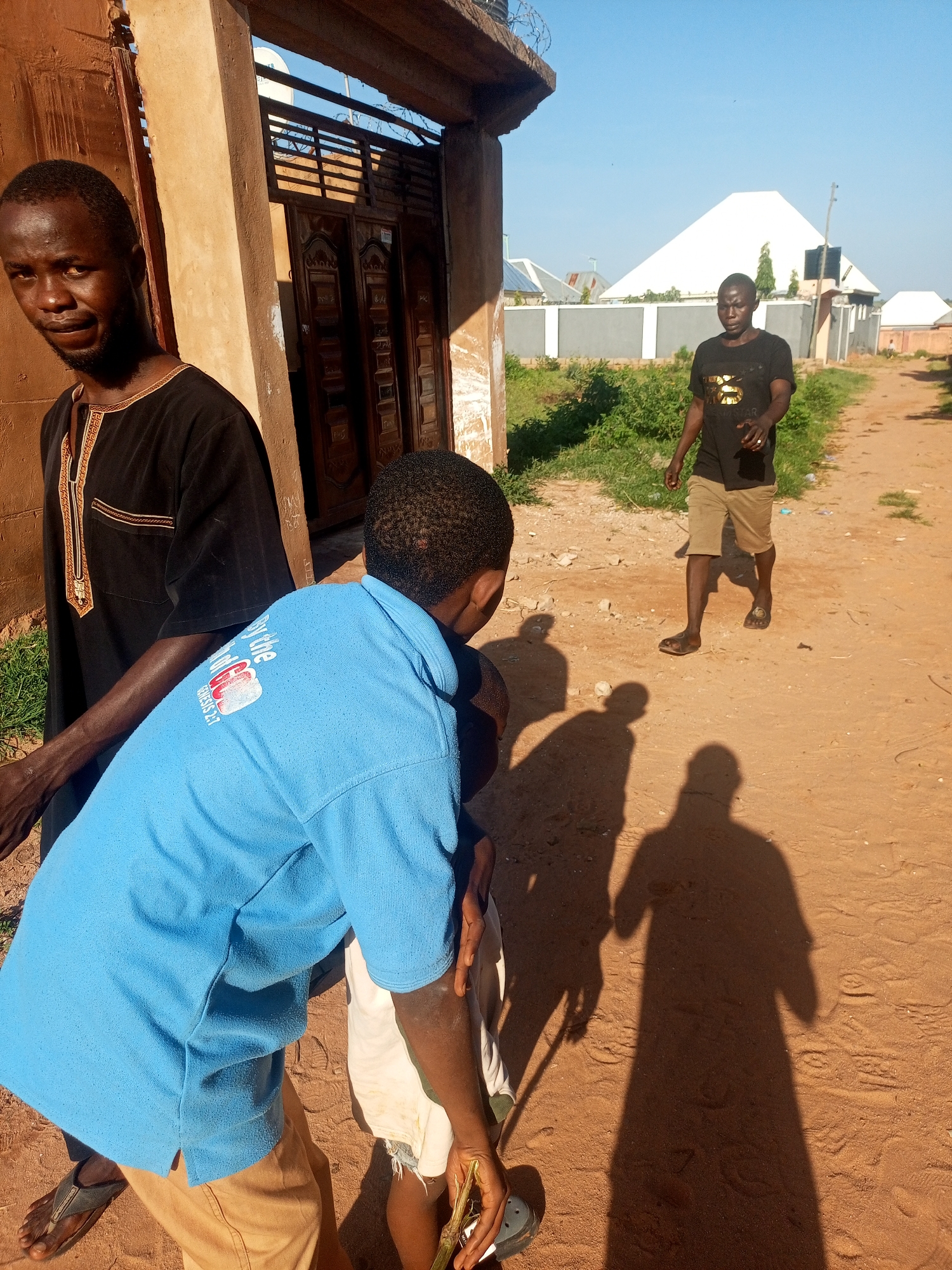 In Tempure, Nigeria, on a sandy path under a clear blue sky, three individuals are present. The foremost subject, a young boy wearing a blue t-shirt with a white graphic print, is partially visible from behind and to the side. He is wearing khaki shorts and dark sandals. To his left stands an adult male in a black traditional outfit with gold trim. His gaze is directed toward the camera. Further down the path, a second adult male, dressed in a black t-shirt with gold lettering and light brown shorts, is walking away from the camera. He is wearing dark sandals and appears to be mid-stride. In the background, a white, pyramid-shaped structure is visible, along with other buildings and vegetation. The ground is a mixture of sand and dirt, with scattered pebbles and debris. The scene is illuminated by sunlight, casting long shadows on the ground.