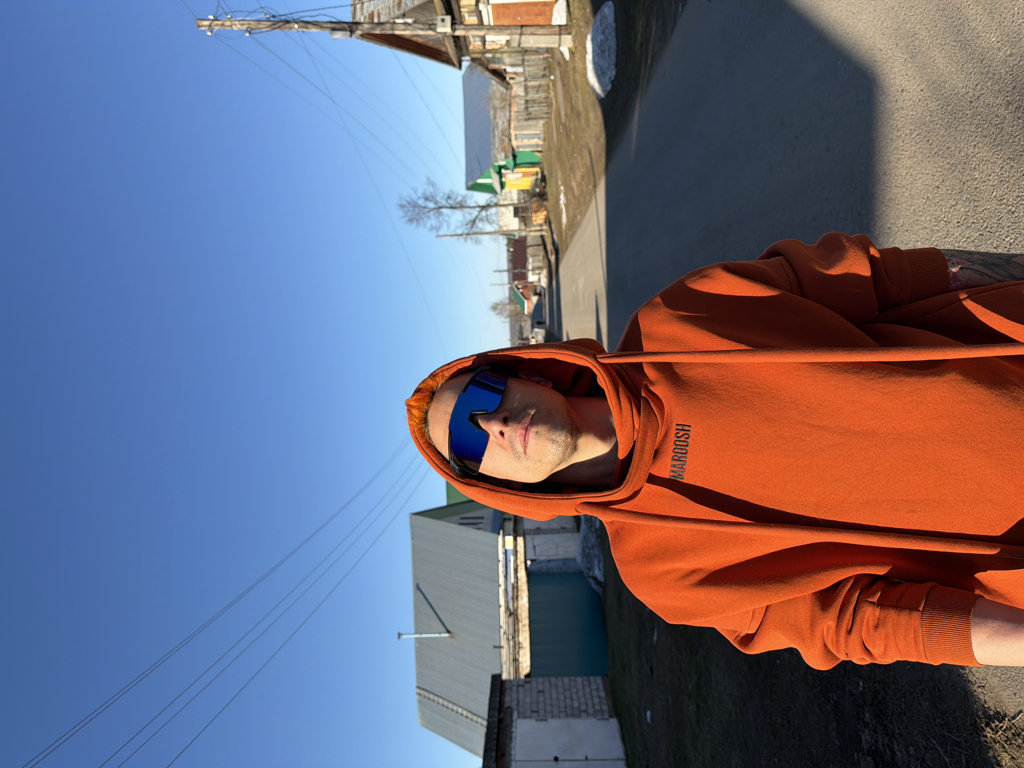 A man wearing an orange hoodie and blue sunglasses is standing on a street in Chaikovsky, Russia. The street is paved and lined with buildings, including houses and a green and yellow structure. A power pole with wires is visible against a clear blue sky. The man is wearing a hoodie with the word "MAROOSH" printed on it. The background includes a mix of residential and urban elements, with signs of early spring such as snow patches and bare trees.