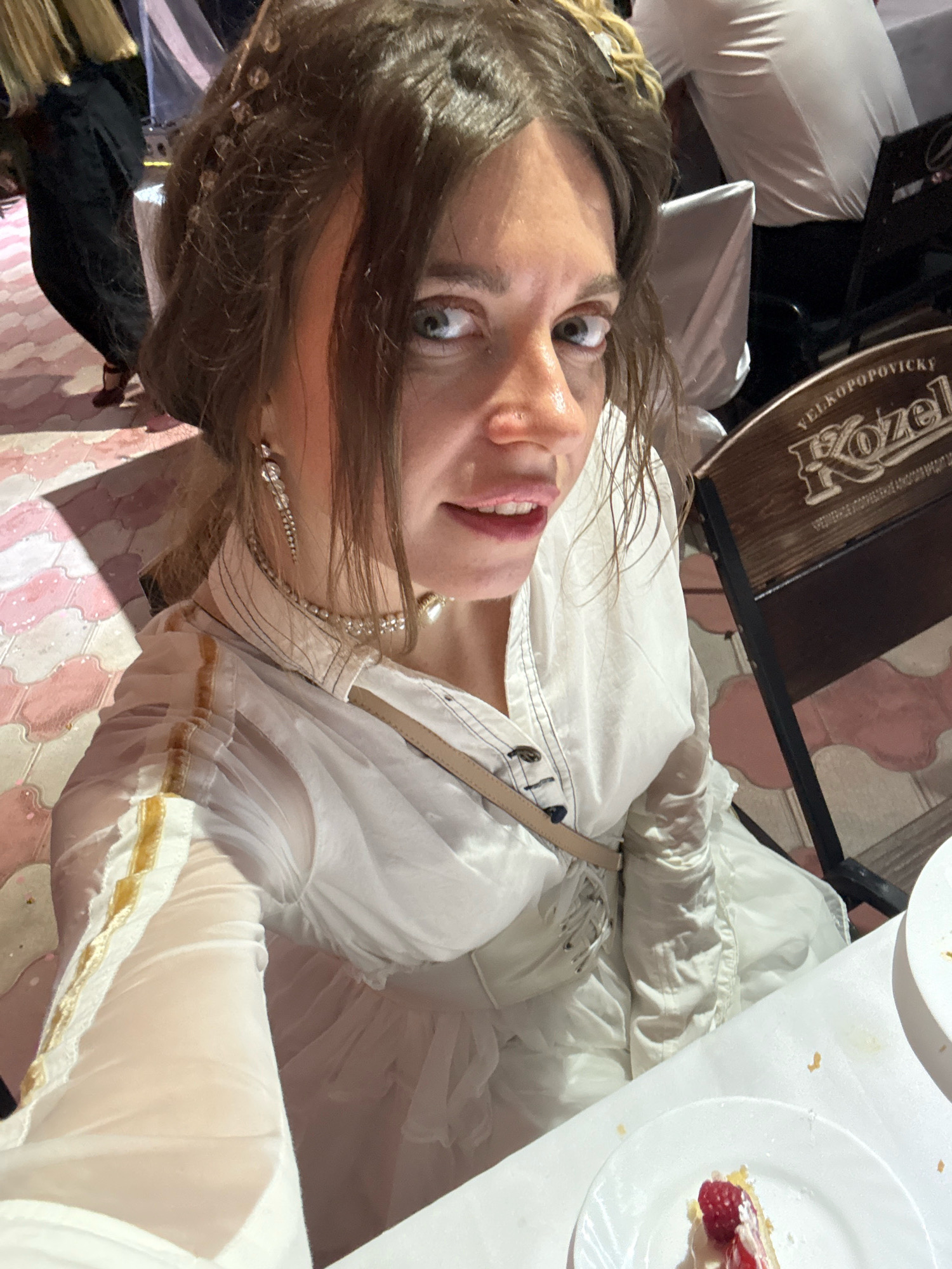The image shows a close-up of a young woman with light brown hair and light green eyes, looking directly at the camera. She is wearing a white, sheer, long-sleeved dress with gold trim on the sleeves and a corset-style lacing detail on the bodice. She has on large, dangling crystal earrings and a delicate necklace. Her makeup appears minimal, with a focus on her lips. The background is out of focus but suggests an outdoor setting with patterned paving stones and hints of other people and structures. To the right of the woman, a wooden chair with the logo "VELKOPOPOVICKY KOZEL" is visible. A white tablecloth is in the foreground, with a slice of cake topped with raspberries and cream on a white plate. The scene is well-lit, possibly by natural light or a warm artificial light source. The woman's hair is styled with intricate braids and small embellishments. The image appears to be a selfie taken at a social gathering or restaurant, with no specific identifiable landmarks of Пакли, Russia.