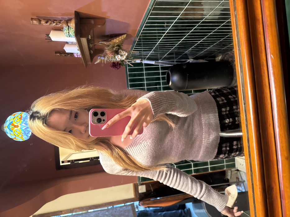 An East Asian female is depicted in a mirror selfie taken within a bathroom environment located in Yuzhong District, China. The subject, visible from mid-thigh up, has long blonde hair with darker roots and wears a light grey V-neck sweater and a dark plaid skirt. She holds a pink-cased smartphone in her right hand, positioned to capture the reflection and partially obscuring her face. Her nails are white with small dark speckles. Her left hand rests near the sink counter, holding a light-colored object. The bathroom features reddish-brown painted upper walls, dark green vertically-oriented subway tiles on the lower walls, and a large mirror with a warm-toned wooden frame. A multi-colored, stained-glass-style dome light fixture is reflected above the subject's head. To the right of the subject, a wooden wall shelf is mounted, displaying two white pillar candles, a green patterned box, and a carved wooden ornament. Beneath the shelf, a dried floral arrangement hangs. A black cylindrical waste bin with a recessed lid is positioned on the tiled floor to the right. A chrome faucet is visible in the reflection at the bottom center. A blurred reflection of another individual or objects is visible to the far left.