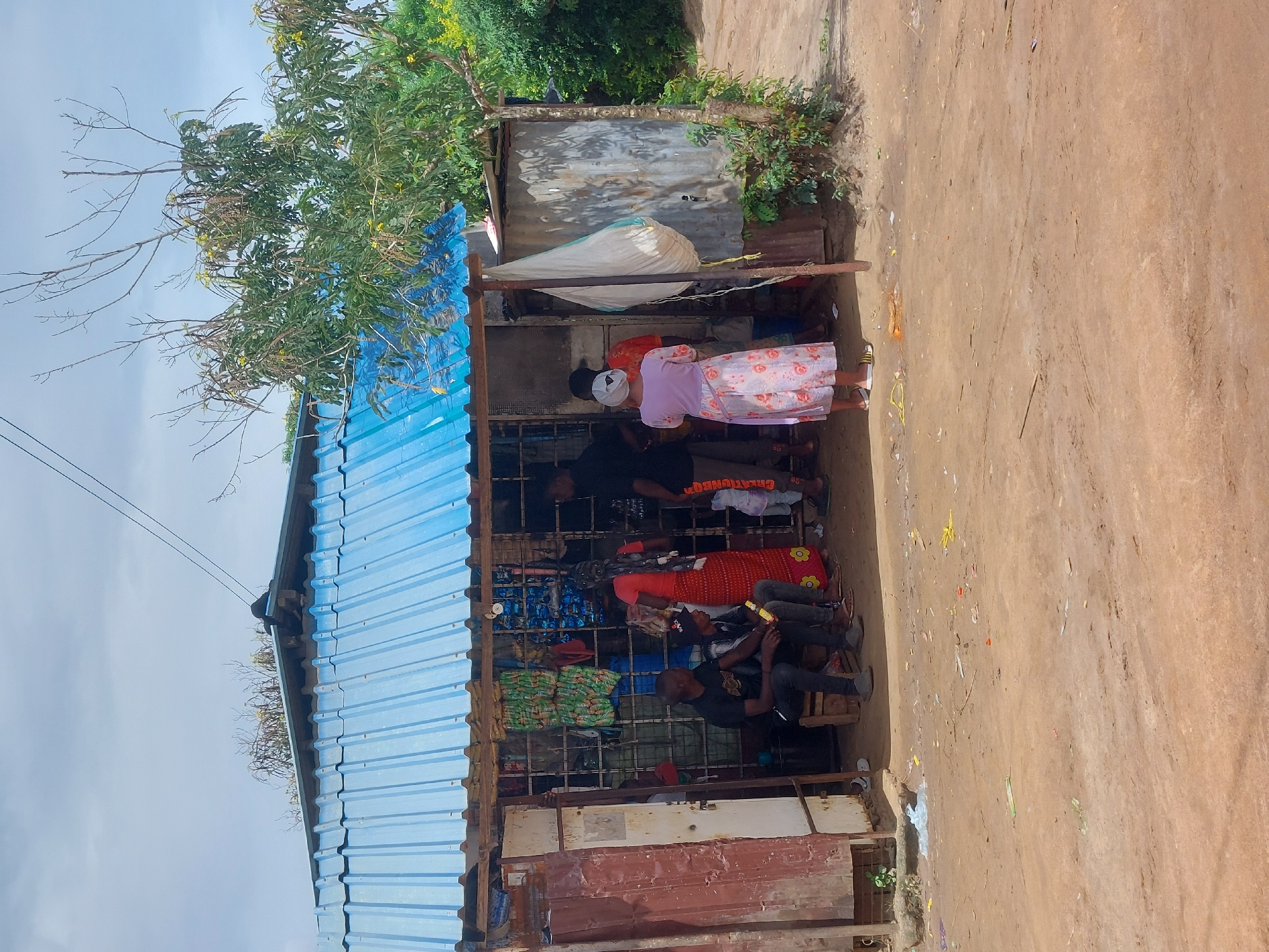 A street-side shop in Dar es-Salaam, Tanzania, is sheltered by a blue corrugated metal roof. The shop's structure is makeshift, utilizing metal bars and corrugated sheeting. Inside, shelves are stocked with various items, including bottled drinks, packaged goods, and clothing. Several people are present. Some are customers standing inside the shop, while others appear to be engaged in conversation. Outside the shop, the ground is bare dirt. Trees and foliage surround the shop. Overhead electrical wires are visible.