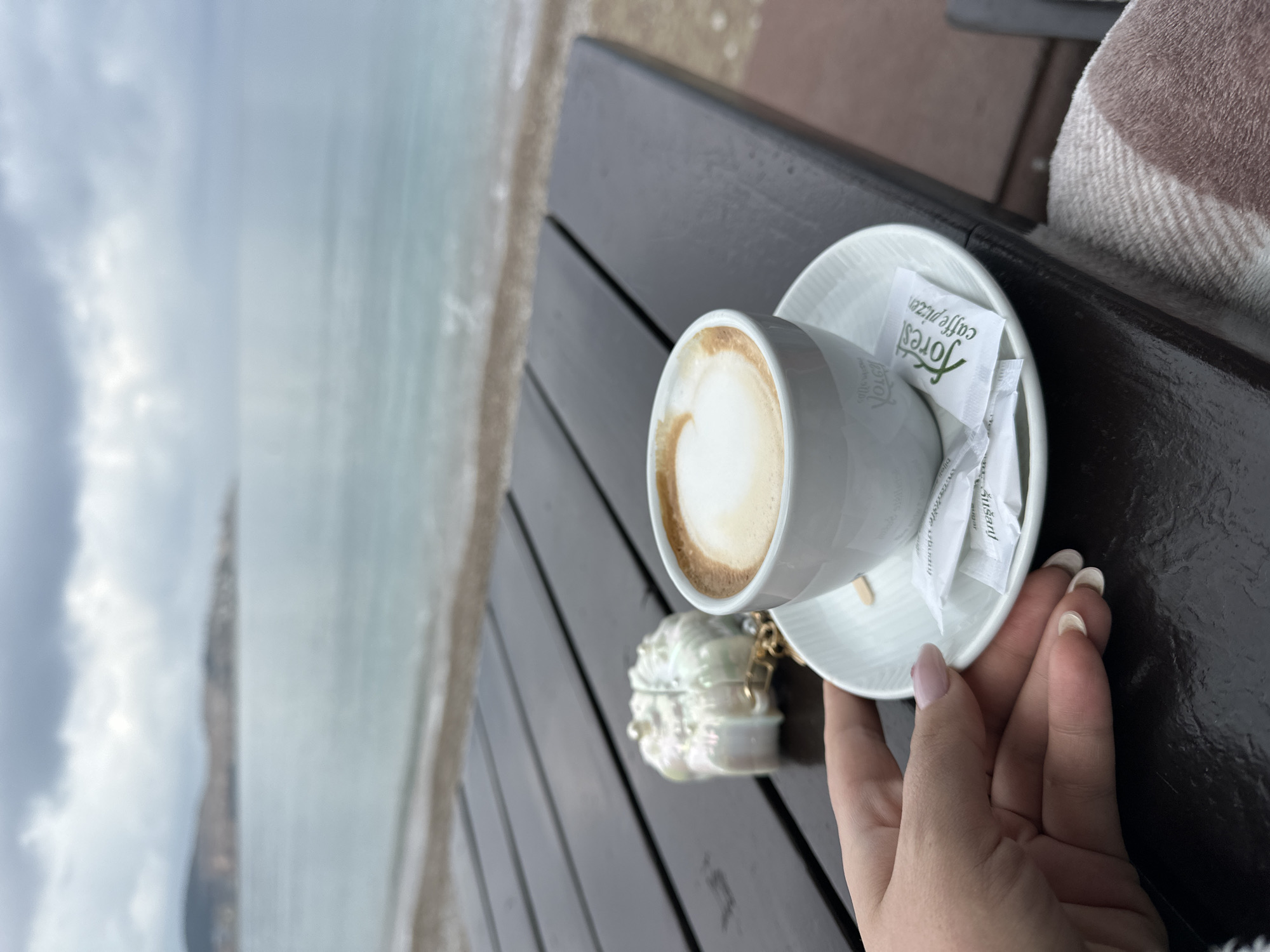 The image shows a cup of coffee on a dark wooden table. The cup is white and sits on a white saucer with sugar packets. A small, ornate box with pearls and gold accents is nearby. A woman's hand rests near the cup. The background features a blurred view of the coast. It is Shushanj, Montenegro. The coast of the city is visible. The sky is overcast.