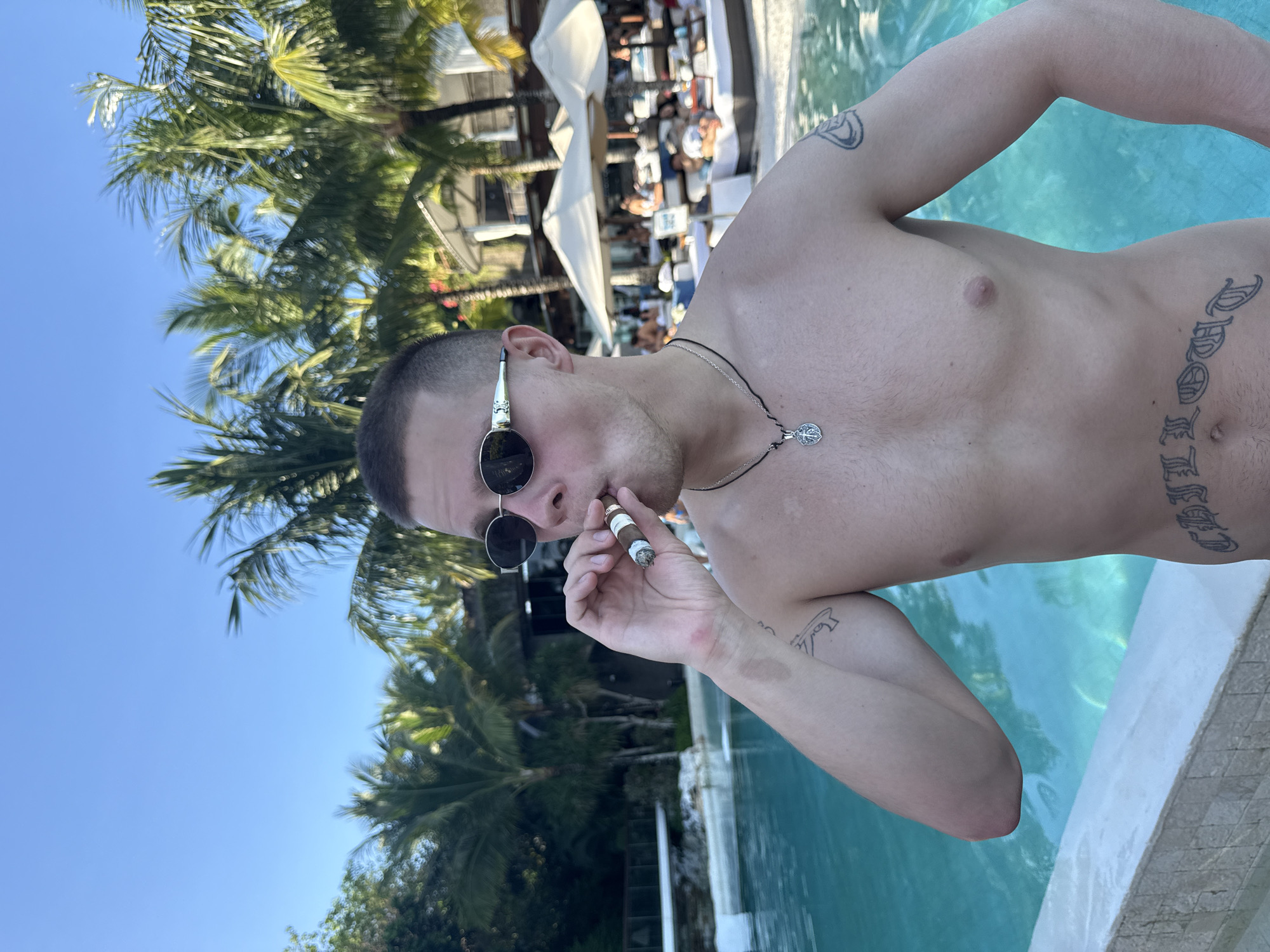 A fair-skinned man stands in a pool, shirtless and wearing sunglasses, smoking a cigar. The scene appears to be set outdoors at a resort or upscale pool area, evidenced by the clear blue water, palm trees, and sun loungers with white umbrellas in the background. The man has short hair and wears a necklace with a pendant. He has multiple tattoos on his arms and torso. The location is Kuta Selatan, Indonesia, based on the tropical vegetation and general resort atmosphere.