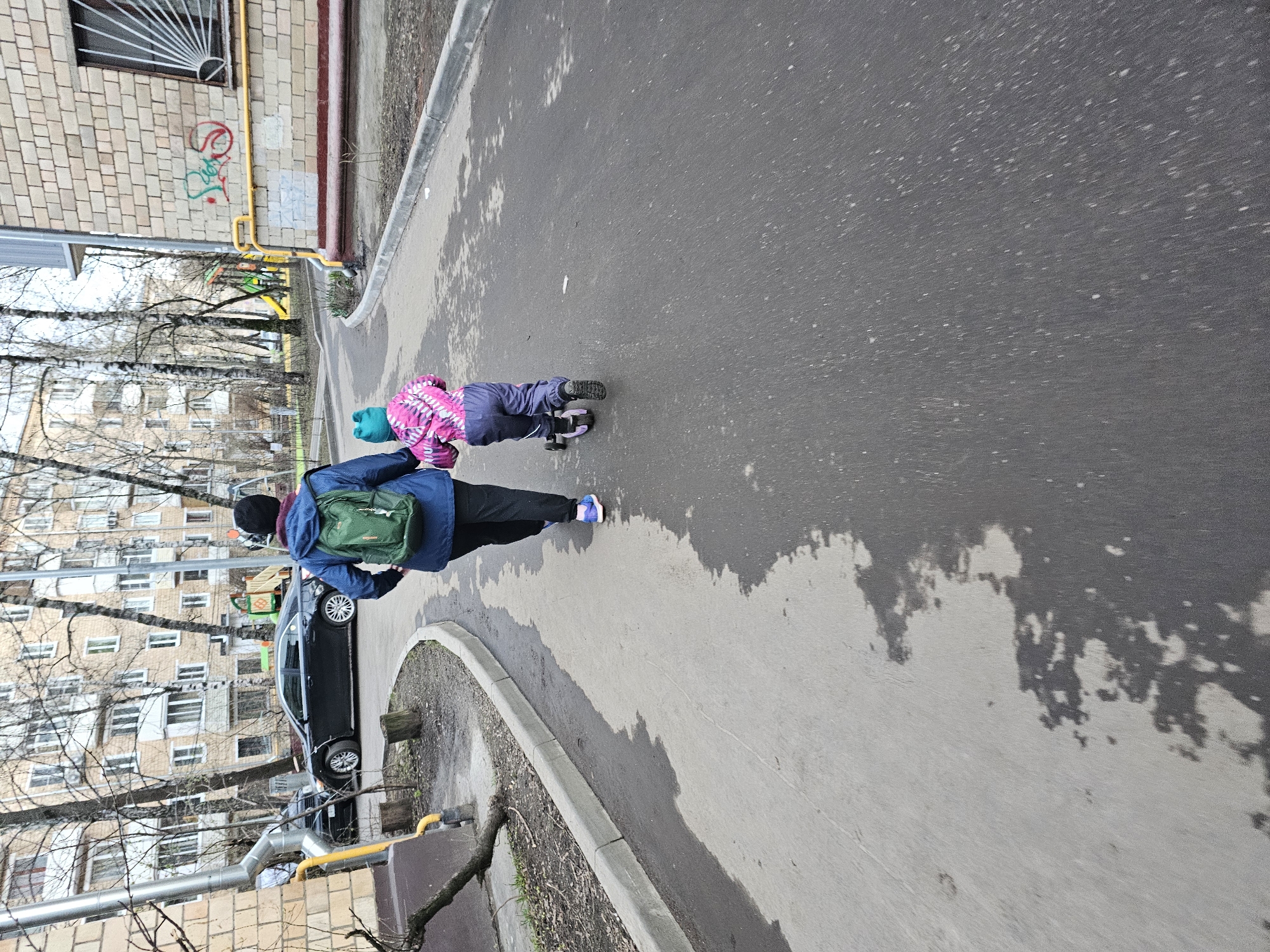 The image depicts a street scene in Khimki, Russia, where an adult wearing a blue jacket, black pants, and a green backpack is walking alongside a child riding a scooter. The child is wearing a pink and purple outfit with a blue hat. They are on a paved surface beside a curb. A black car is parked nearby. Tall apartment buildings and trees are visible in the background. The lighting suggests a cloudy day. The ground shows signs of dampness or shadows.