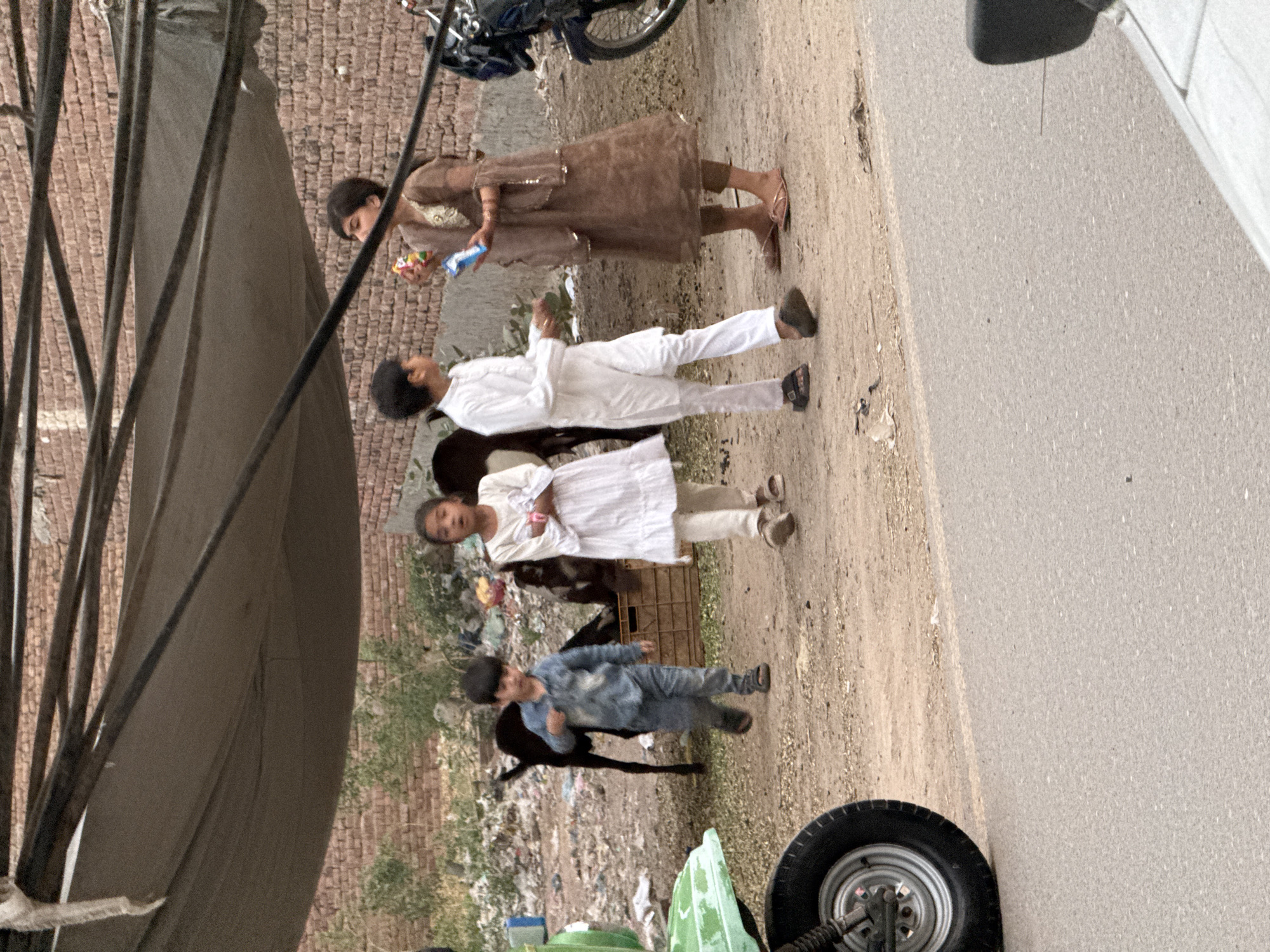 The image shows a dirt path on the right and a building on the left. A tent or tarp is partially visible on the left side of the building. There is a group of children on the path, dressed in various clothes. A young girl in a brown outfit is at the top of the group, followed by a child in white. Another child in white is below the second child and another child dressed in a blue outfit is at the bottom. Goats are visible among the children. Trash is seen on the ground to the left. A vehicle wheel is visible at the bottom right corner, and another is partially visible at the top. The location seems to be an undeveloped or impoverished area in a city.