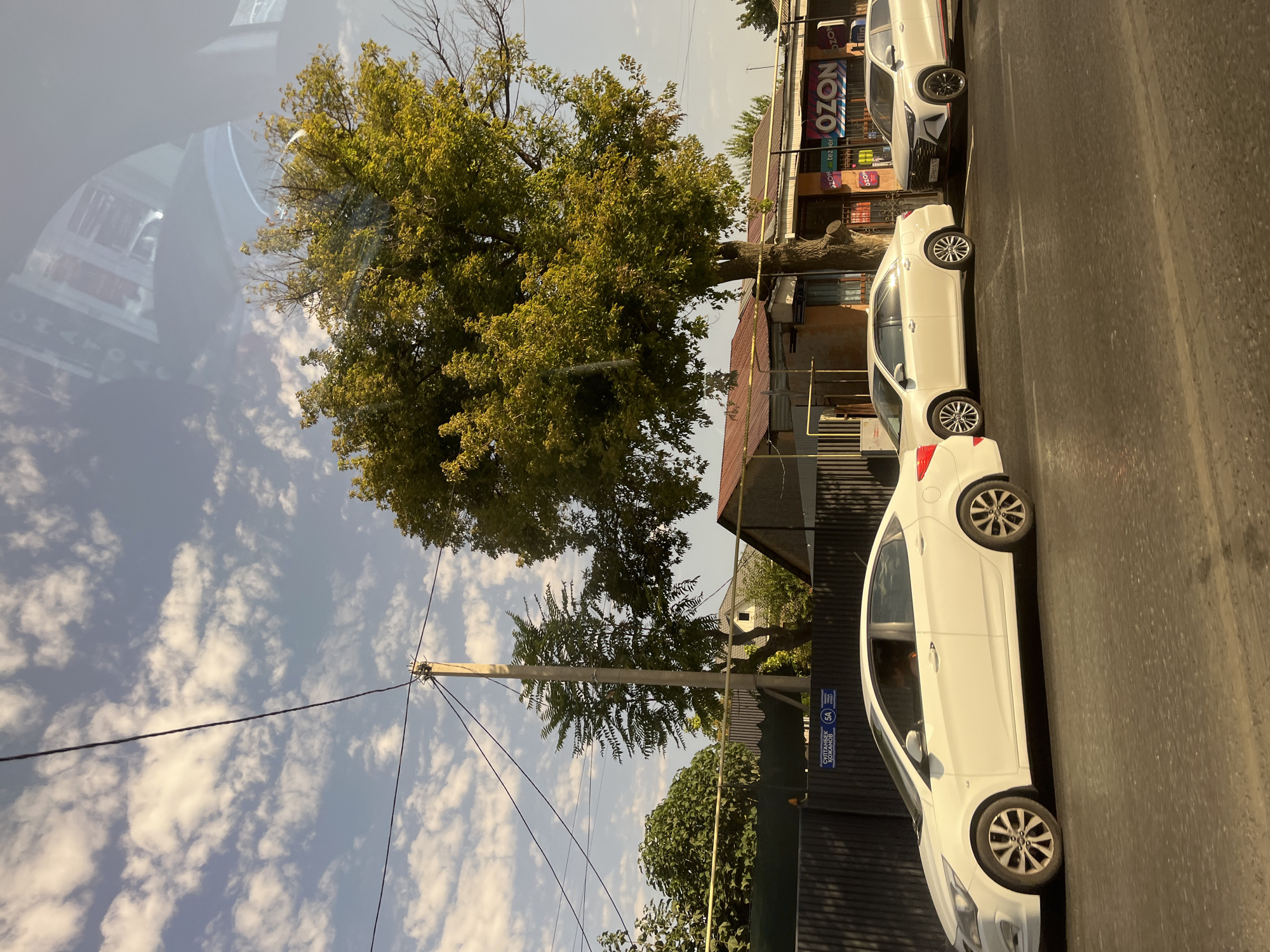 The image captures a street scene in Shymkent, Kazakhstan, under a bright, partly cloudy sky. In the foreground, a white Hyundai sedan is parked on the right side of a paved road. To its right, two other white cars are visible: a sedan further down the street, and a white SUV parked in front of a building. The buildings lining the street appear to be commercial establishments, with one clearly marked with an "OZON" logo, a Russian online retailer. A large tree with green foliage dominates the mid-ground, casting shadows on the street and buildings. Several power lines are strung across the sky, supported by a concrete pole. A dark metal fence runs along the property line behind the parked cars, and glimpses of houses and greenery are visible beyond it. The overall impression is a typical urban street on a sunny day.