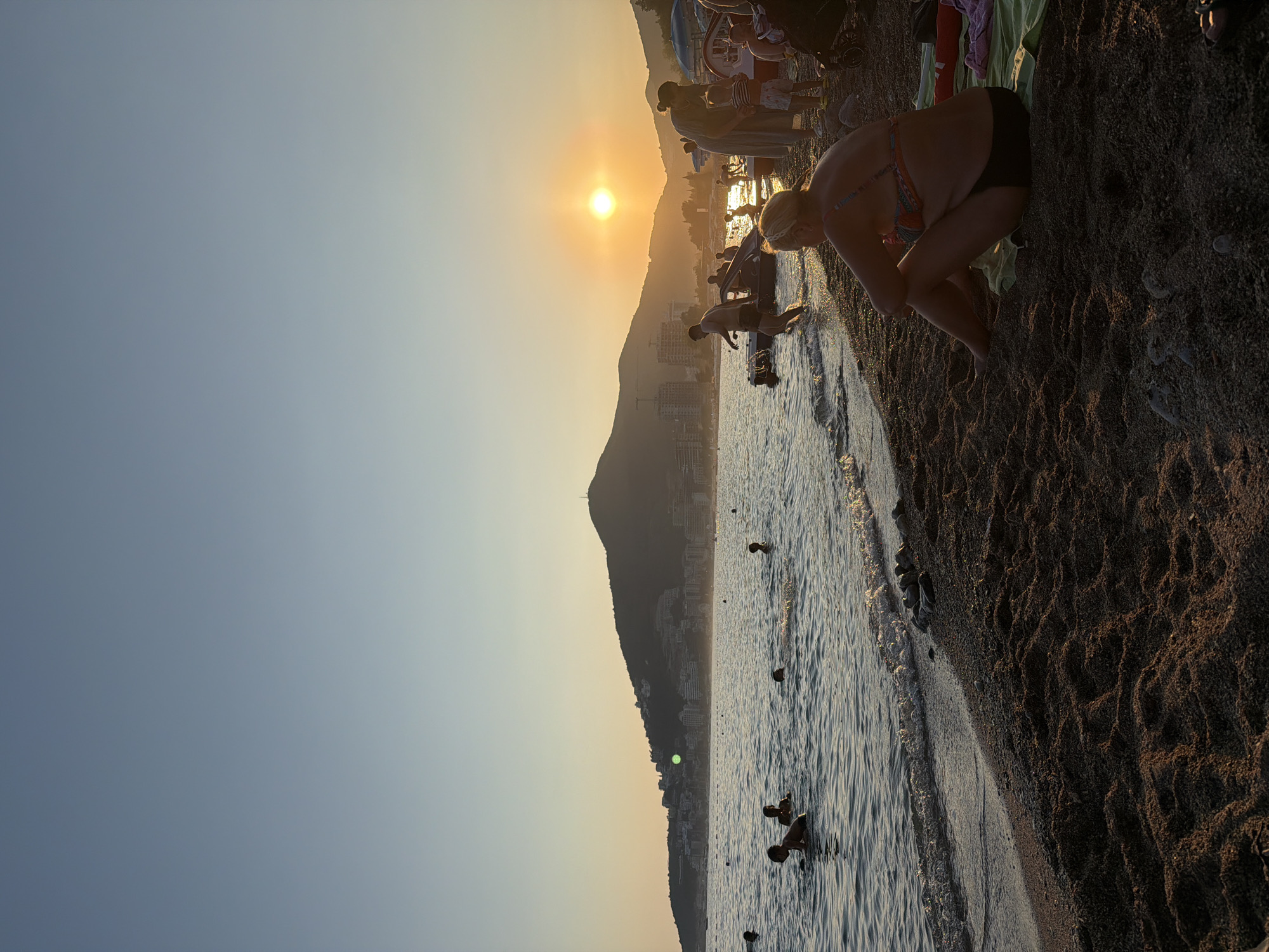 A wide-angle, high-angle shot captures a beach scene in Bečići, Montenegro, during sunset. The sky transitions from a clear, light blue at the top to a warm orange hue near the horizon, where the sun is setting behind a large, dark mountain range. Buildings are visible along the mountain slopes. The sea is calm with gentle waves lapping at the shore.
Several people are in the water, including children swimming and playing. On the beach, a woman is sitting on a towel facing away from the camera, wearing a bikini. Other people are standing near the water's edge, some by a small boat. Beach umbrellas and a stroller are also present. The beach itself is composed of dark sand and pebbles. The overall impression is a serene evening at a popular tourist destination.