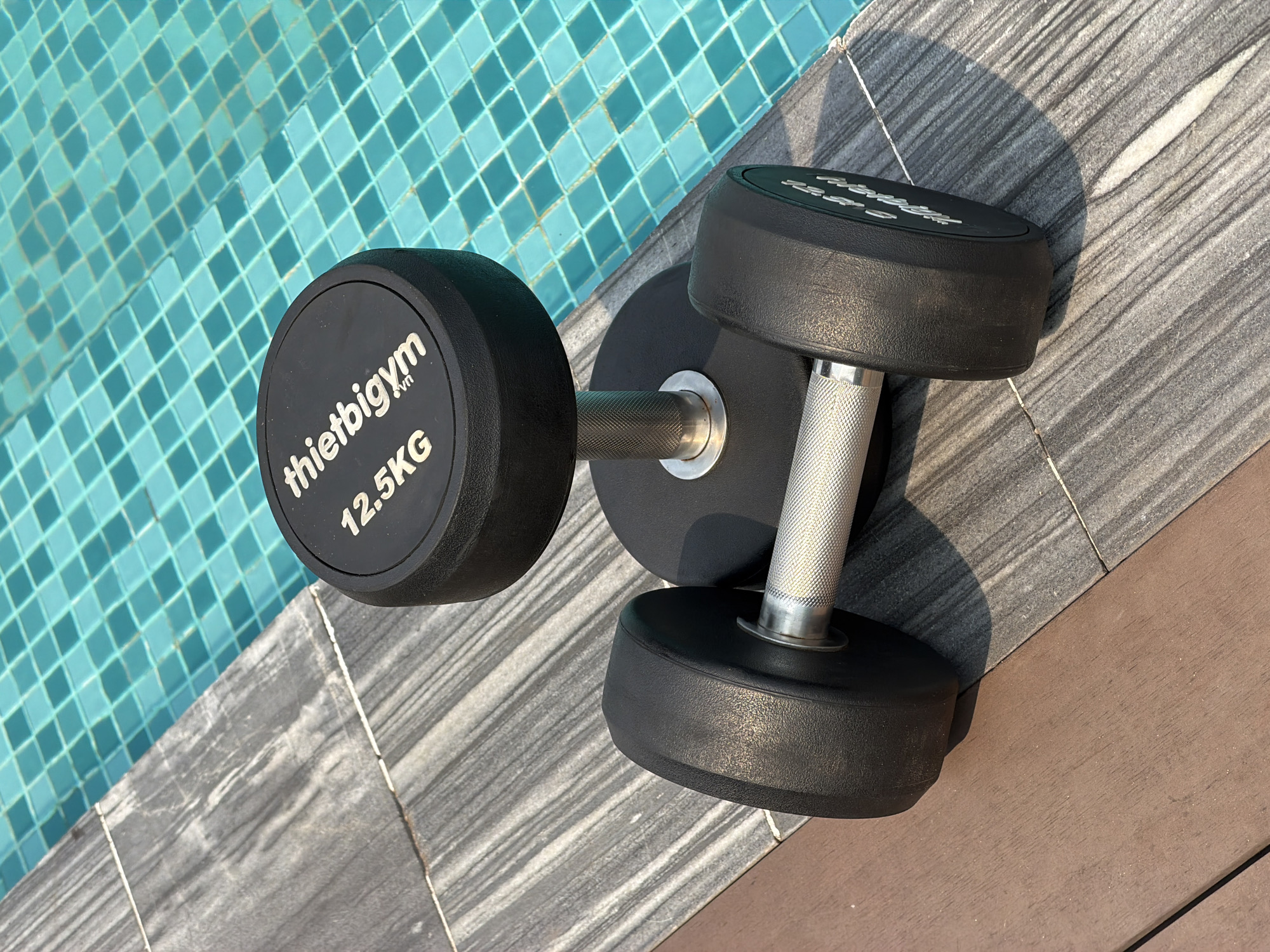 A dumbbell rests on the edge of a swimming pool. One end of the dumbbell sits on grey tile, and the other end is near the pool's turquoise tile. The dumbbell has black weight plates labeled "thietbigym.vn 12.5KG," and a silver knurled handle. The floor transitions from grey tile to brown wood-like material. The scene is in daylight. The presence of the weight in close proximity to the pool could suggest the location is near the water, inside a gym or a private residence. Given the branding on the dumbbell, "thietbigym.vn", and the .vn domain, it is most likely that the location is in Vietnam. Đà Nẵng is suggested by the website.
