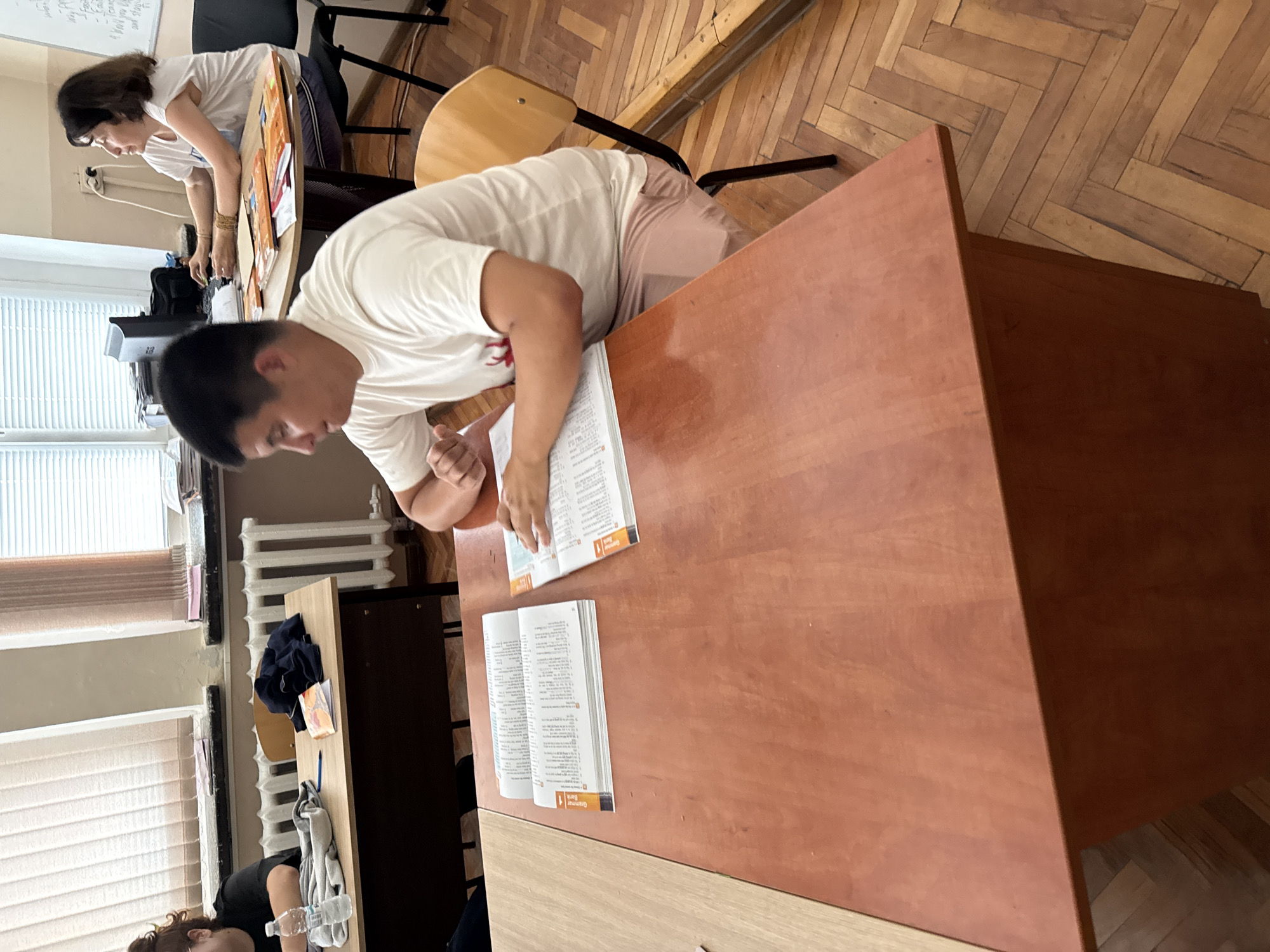 The image depicts a classroom scene in Elhovo, Bulgaria. A young man is bending over a wooden desk, apparently studying from a textbook. A book is open in front of him, placed on the desk's surface. In the background, another individual, possibly a woman, is leaning over a different desk, engaged in an activity not fully visible. The room is illuminated by natural light coming through the windows to the side. The environment includes typical classroom elements such as desks, chairs, and a radiator. Wooden floors are visible, adding to the scene's background.
