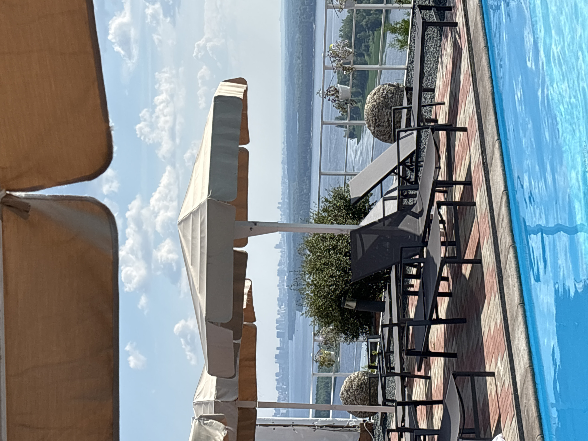 The image depicts a scenic view of a river, with a city skyline in the distance, as seen from a poolside deck in Novokuybyshevsk, Russia. The foreground features a bright blue swimming pool, bordered by a gray coping stone. Beyond the pool, a paved patio area is furnished with several dark gray lounge chairs and small tables, positioned under two large, light-colored umbrellas that provide shade. A white metal railing separates the patio from a vegetated area with trees and shrubs, beyond which the wide river flows. In the background, a distant cityscape with buildings can be observed, silhouetted against a hazy sky with scattered white clouds. The overall impression is one of a tranquil outdoor leisure space overlooking a expansive natural and urban landscape. There are no people visible in the image.