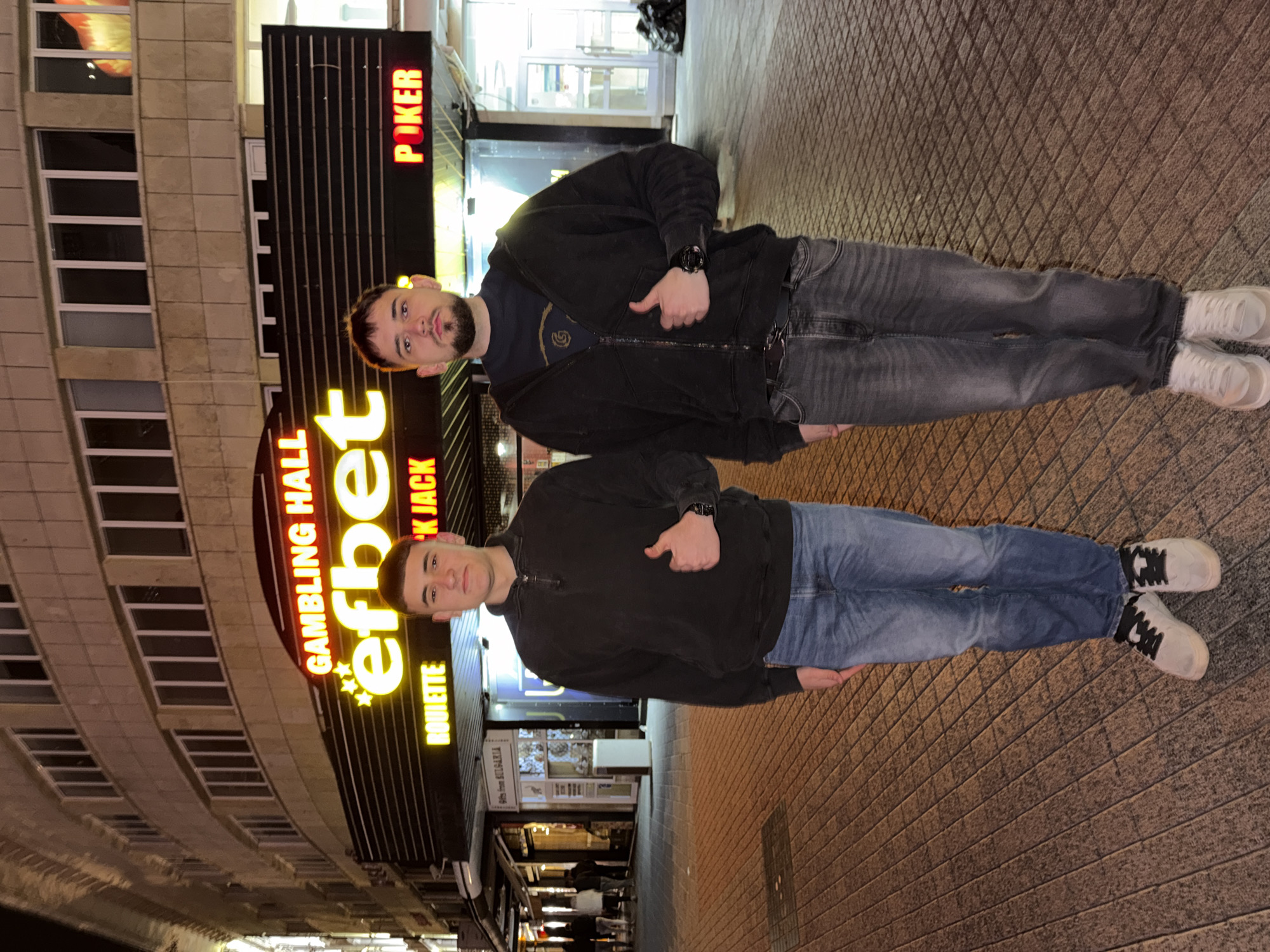Two men are standing on a brick-paved walkway in front of a gambling hall called "efbet". The gambling hall has neon signs for "POKER", "BLACK JACK", and "ROULETTE". The building is multi-story and has many windows. The walkway appears to be in a narrow street, with other businesses visible in the background. The location is in the city of Plovdiv, Bulgaria. Both men are giving a thumbs-up. The first man is wearing a black jacket, a dark blue t-shirt, grey jeans, and white sneakers. The second man is wearing a black sweatshirt, blue jeans, and white sneakers with black laces.