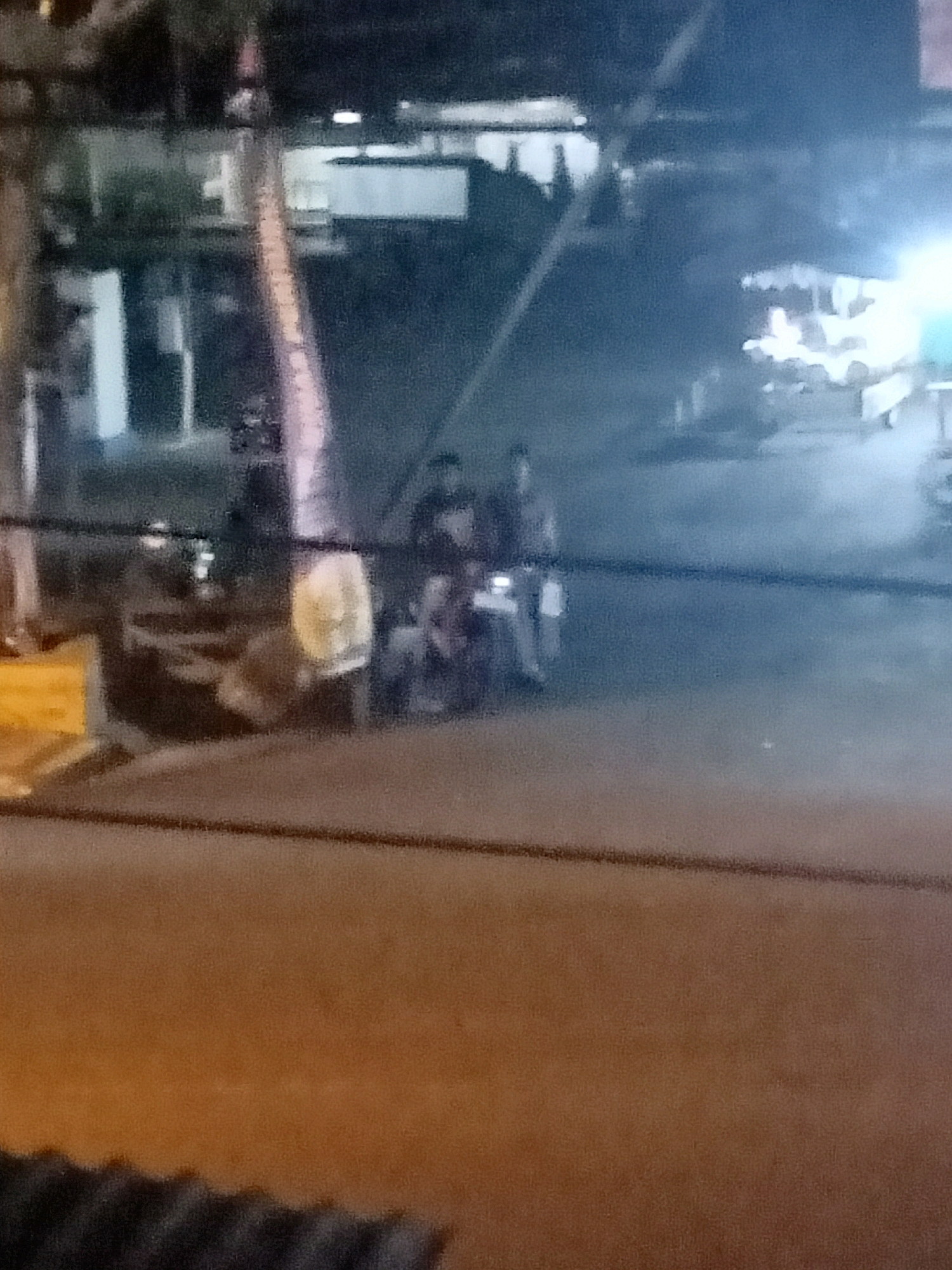 The image depicts a nighttime street scene in Jatisrono, Indonesia. A blurry motorcycle with two occupants is visible in the mid-ground, with the rider appearing to be wearing a helmet and the passenger possibly wearing a white shirt. The motorcycle has a bright yellow headlight. To the left of the motorcycle, a portion of a light-colored, vertical structure, possibly a pole or a thin tree trunk, is visible. The background shows indistinct shapes of buildings and some illuminated areas, suggesting shops or other establishments. The foreground is dominated by a dark, horizontal line across the image, possibly a railing or wire, and a textured, brown surface below it. The overall image quality is low, with significant blur and low light conditions, making precise identification of details challenging. The scene suggests a casual street environment, likely a commercial or residential area at night.