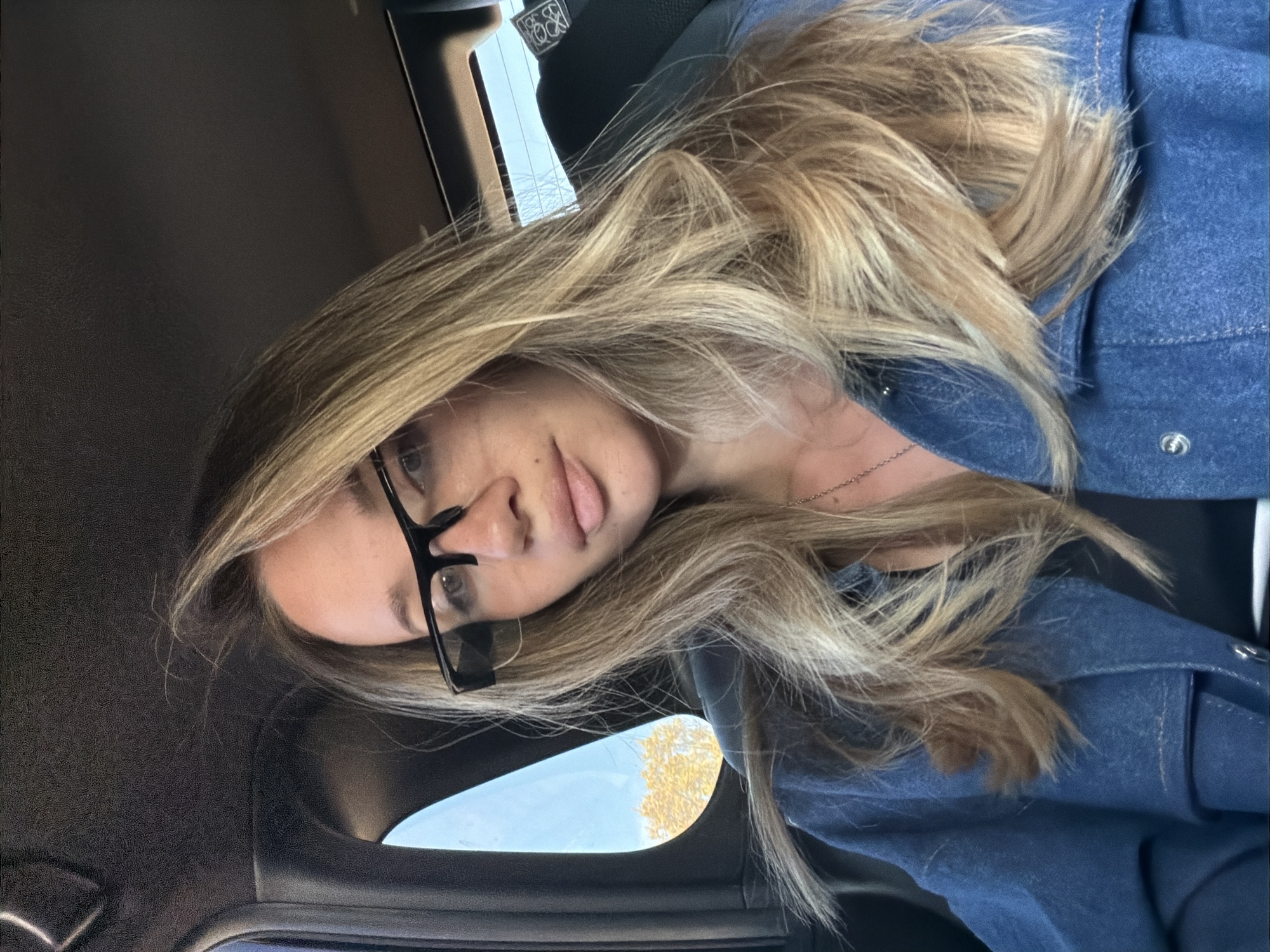 The image shows a woman inside a car. She is wearing glasses and a blue denim jacket. Her hair is long and blonde. Through the car window, a blue sky and a tree with yellow leaves can be seen. The location appears to be inside a car. The city is unknown.