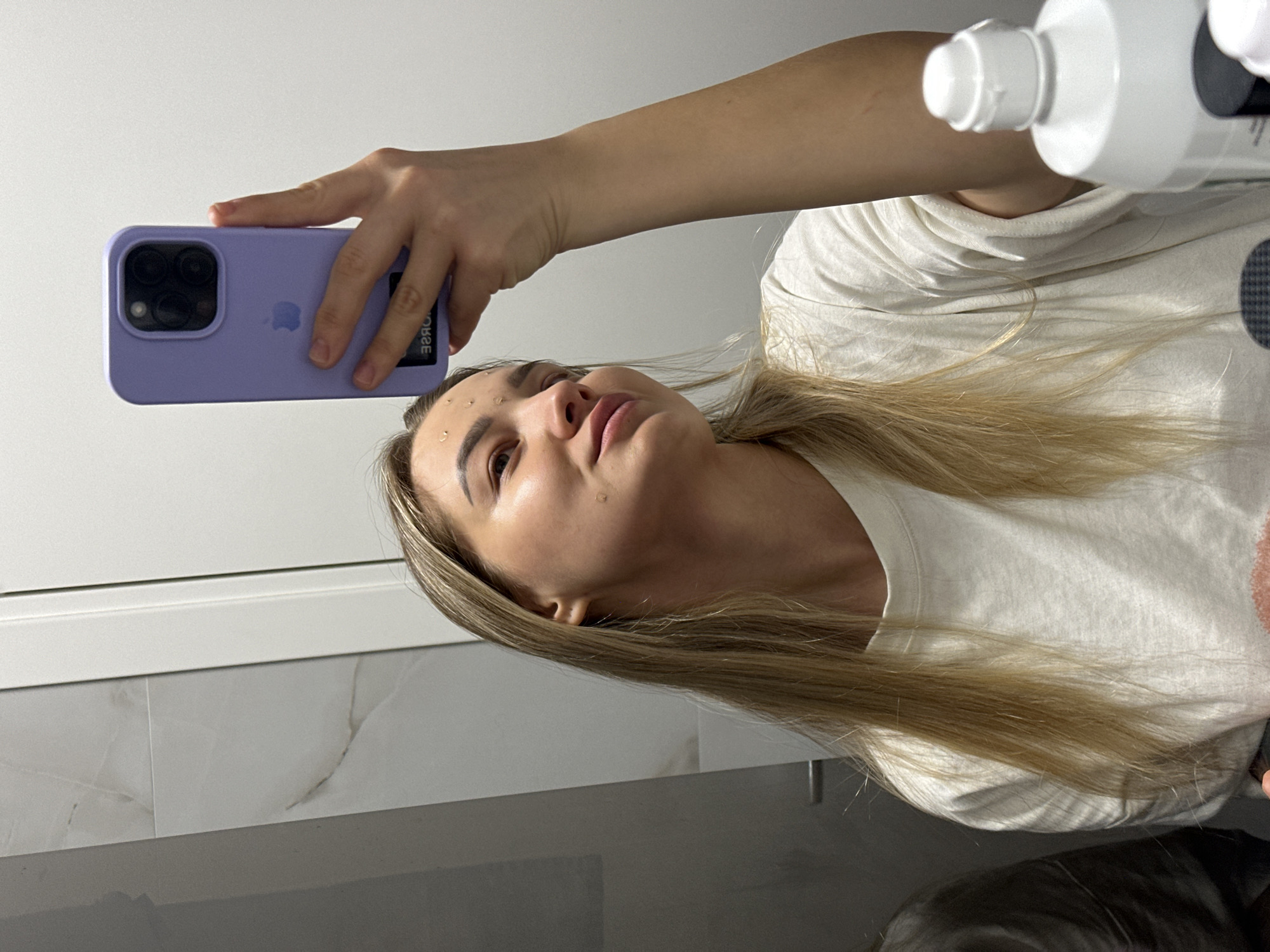 A blonde woman in a white t-shirt takes a selfie in a bathroom. She is holding a purple iPhone in her left hand and a white bottle in her right. There are water droplets on her face. The bathroom has white marble tiles on the wall. The image does not provide enough information to confirm the location is in Budva, Montenegro.