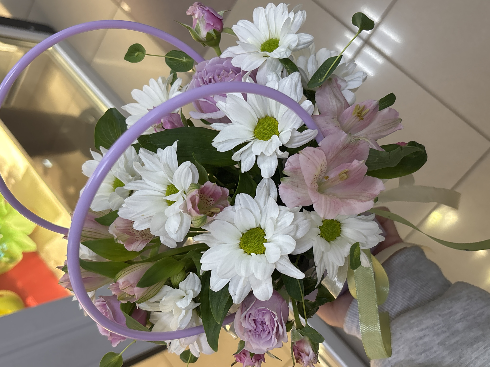 An overhead shot captures a bouquet of white daisies and pink alstroemeria flowers arranged in a light purple basket. The basket has two circular handles and a ribbon tied around its base. The bouquet is being held by a person wearing a grey knitted sweater. The background is blurred, showing a reflective surface with hints of green and red.