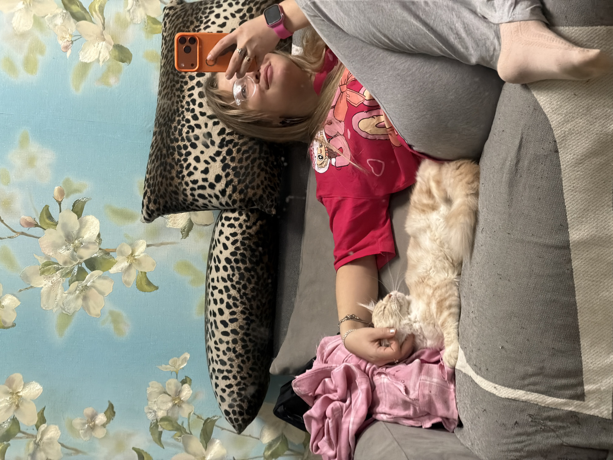 A person with glasses and a pink smartwatch is lying on a gray couch, holding an orange smartphone. They are wearing a red shirt with cartoon characters and gray pants. A fluffy, light orange cat is nestled next to the person on a pink and gray plaid blanket. To the left, there are two patterned pillows against a blue wall decorated with white floral designs. The photograph appears to be taken indoors. There is no information in the image to confirm a location within the city of Chaikovsky, Russia.