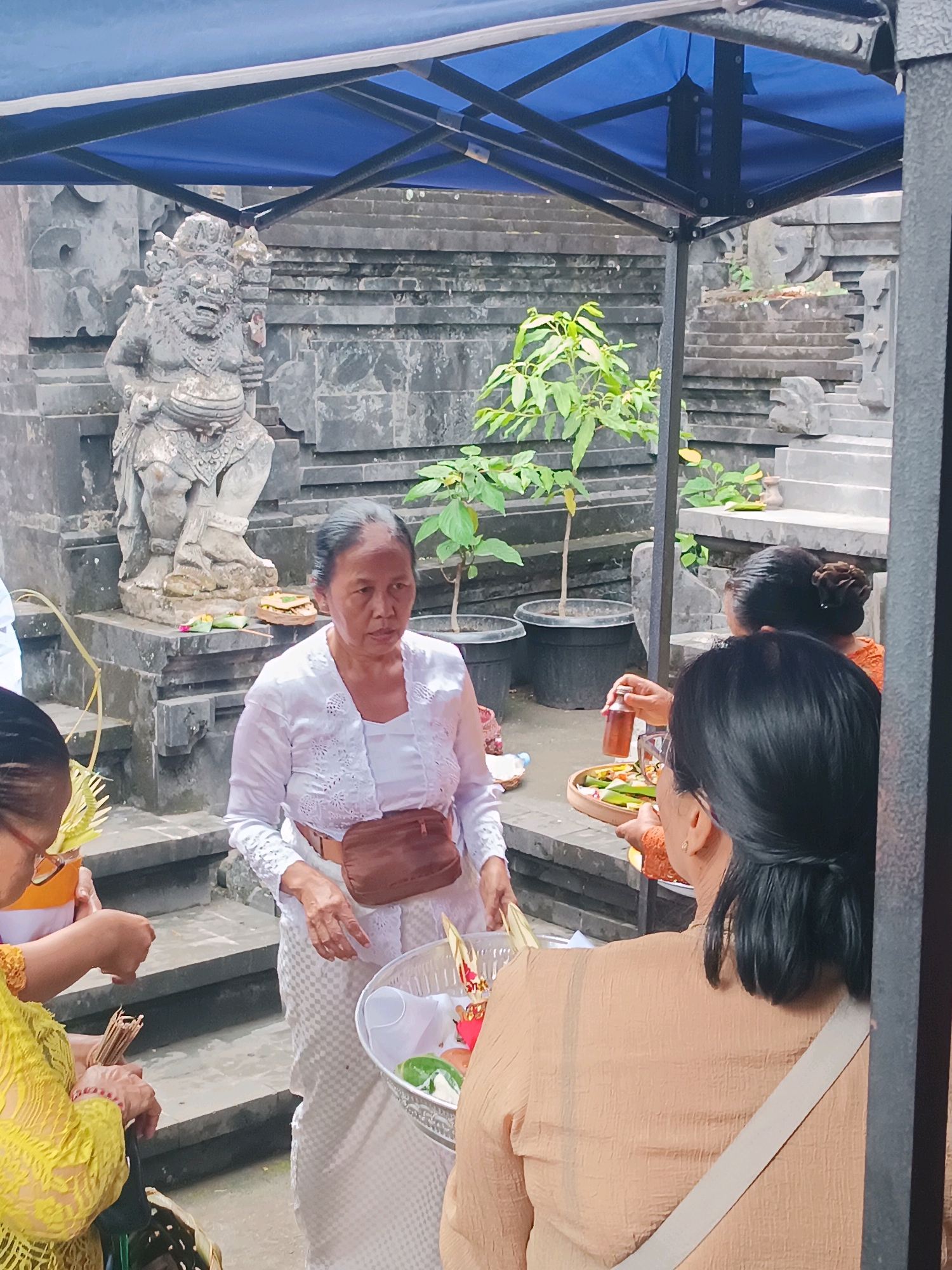 In Tabanan, Indonesia, a woman in a white outfit and a brown fanny pack stands near a stone statue at a temple. She appears to be participating in a religious ceremony. In front of her, a person in a yellow shirt is holding a bundle of sticks, while another person in an orange shirt offers a bottle and a basket filled with offerings. In the background, stone steps and lush greenery suggest the serene and spiritual atmosphere of the temple grounds.