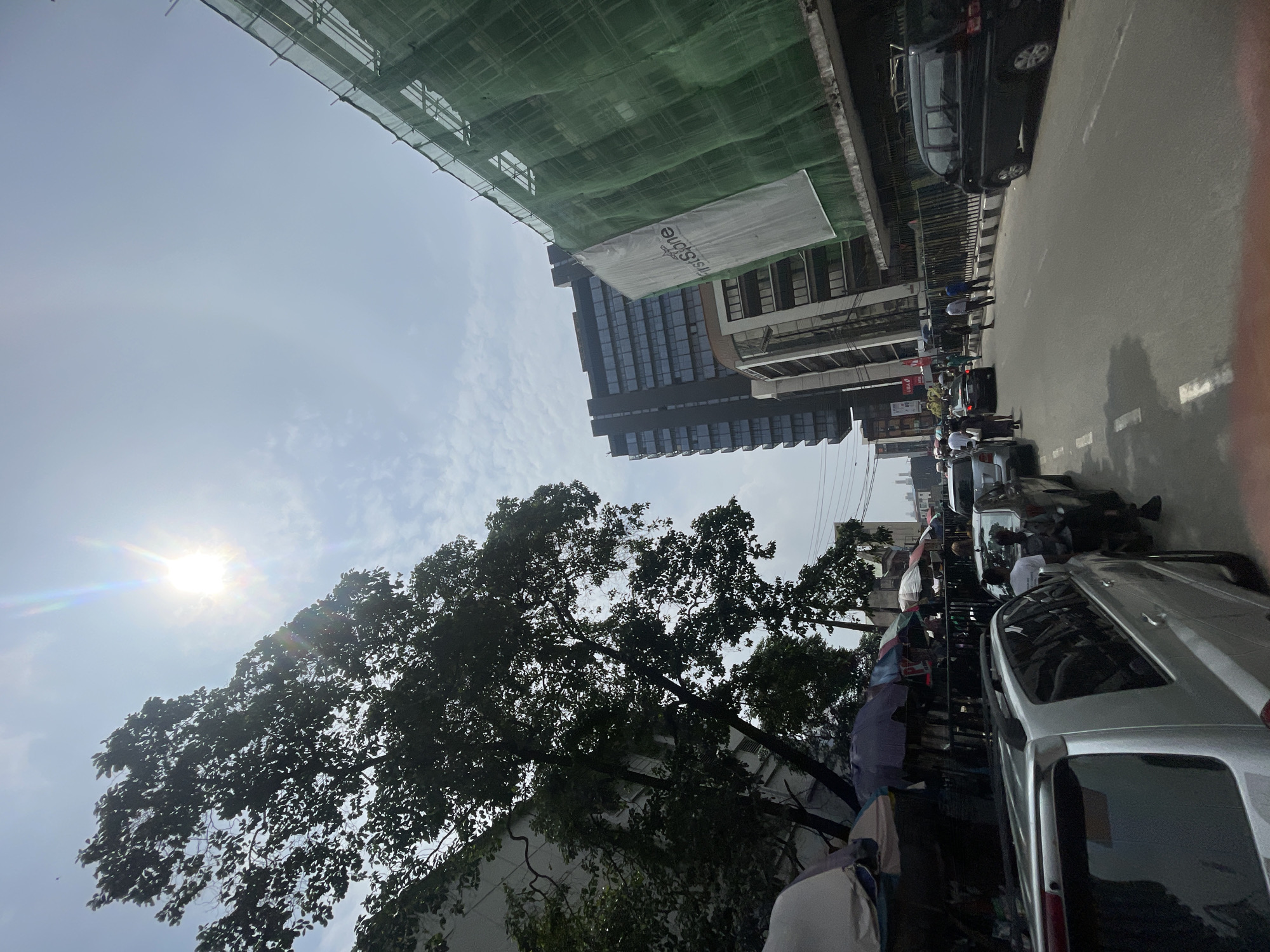 The image depicts a street scene in Lagos, Nigeria. The shot is taken from a low angle, looking upwards.
In the foreground, there's a partially visible silver SUV parked on the right side of the frame. Pedestrians are walking on the side of the road, some appearing to be vendors with umbrellas. A line of vehicles stretches into the distance, indicating traffic.
On the left, there's a large tree with dense foliage partially obscuring a building. Above the tree, the sun is visible, causing a lens flare effect. Further up, there are buildings under construction, covered with green scaffolding and netting. A white banner with text is attached to one of the buildings. In the background, taller buildings are visible against a light blue sky.